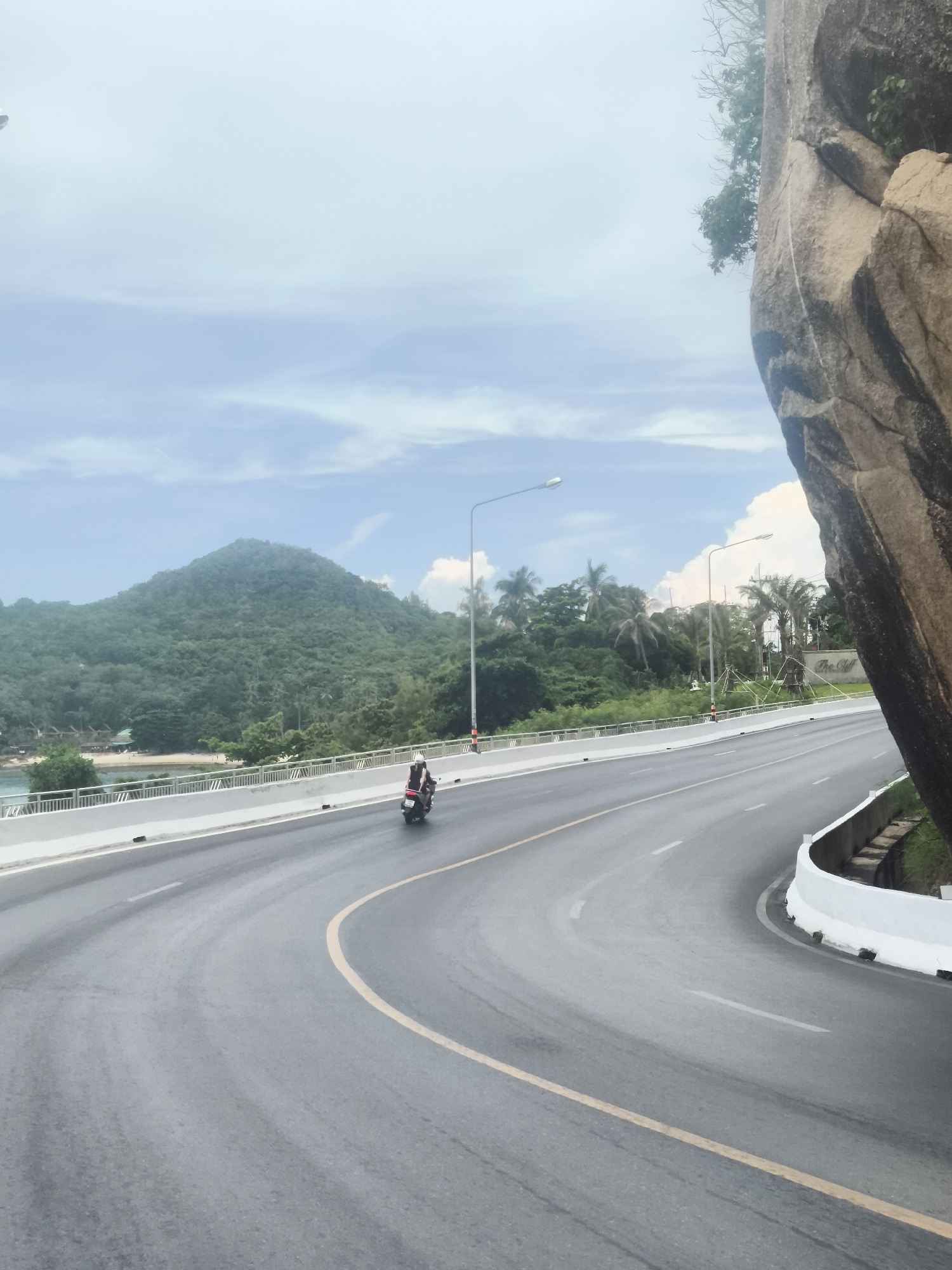 The image captures a scenic view of a winding road along a coastline, with a large rock formation on the right and a lush green mountain in the background. The road appears to be a multi-lane highway with distinct yellow and white lane markings. A lone motorcyclist is visible on the road, riding a scooter away from the viewer. The environment is characterized by tropical vegetation, including palm trees and dense foliage on the mountain. The sky is cloudy, with patches of blue visible. In the distance, there are buildings or structures nestled near the coastline, hinting at a resort or residential area. A sign that reads "The Cliff" is partially visible on the right side of the road, suggesting the name of a resort or establishment. The overall impression is one of a beautiful, coastal drive in a tropical setting. The location is identified as Baan Chaweng Noi, Thailand.
