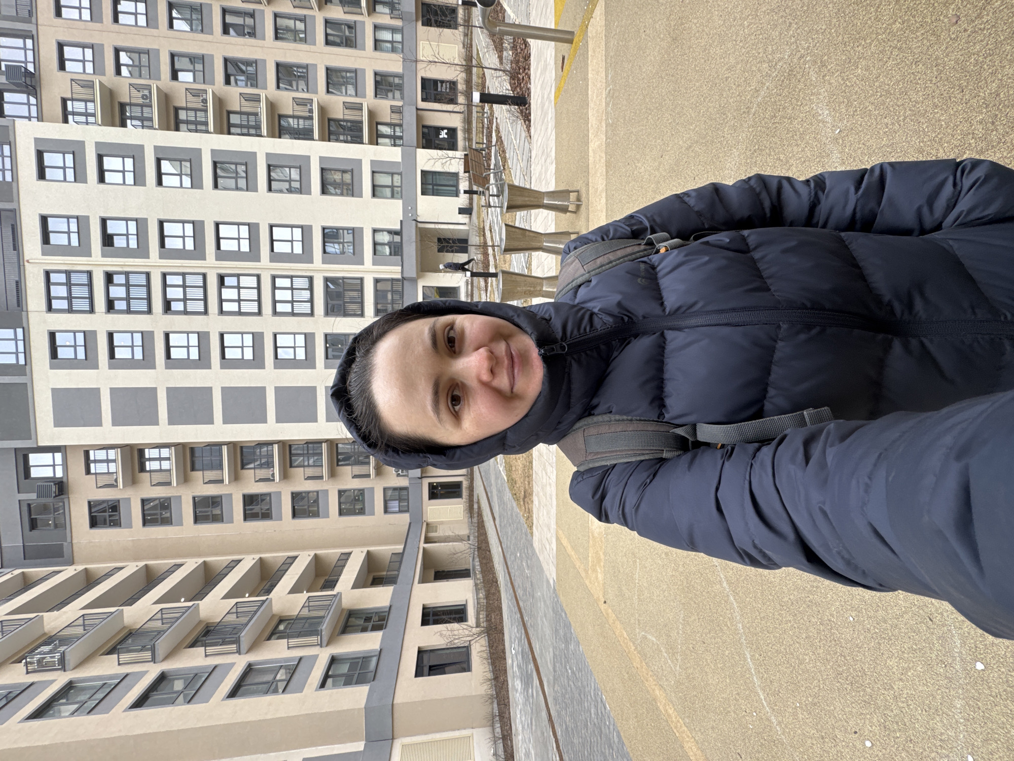 A woman is taking a selfie outside an apartment building in Izhevsk, Russia. She is wearing a dark blue hooded puffer jacket and a gray backpack. Behind her is a tall apartment building with multiple floors and rows of windows and balconies. In front of the building are metal tables and benches. The ground is a mix of pavement and a yellowish surface. The photo is taken at an angle, causing the background to appear tilted.