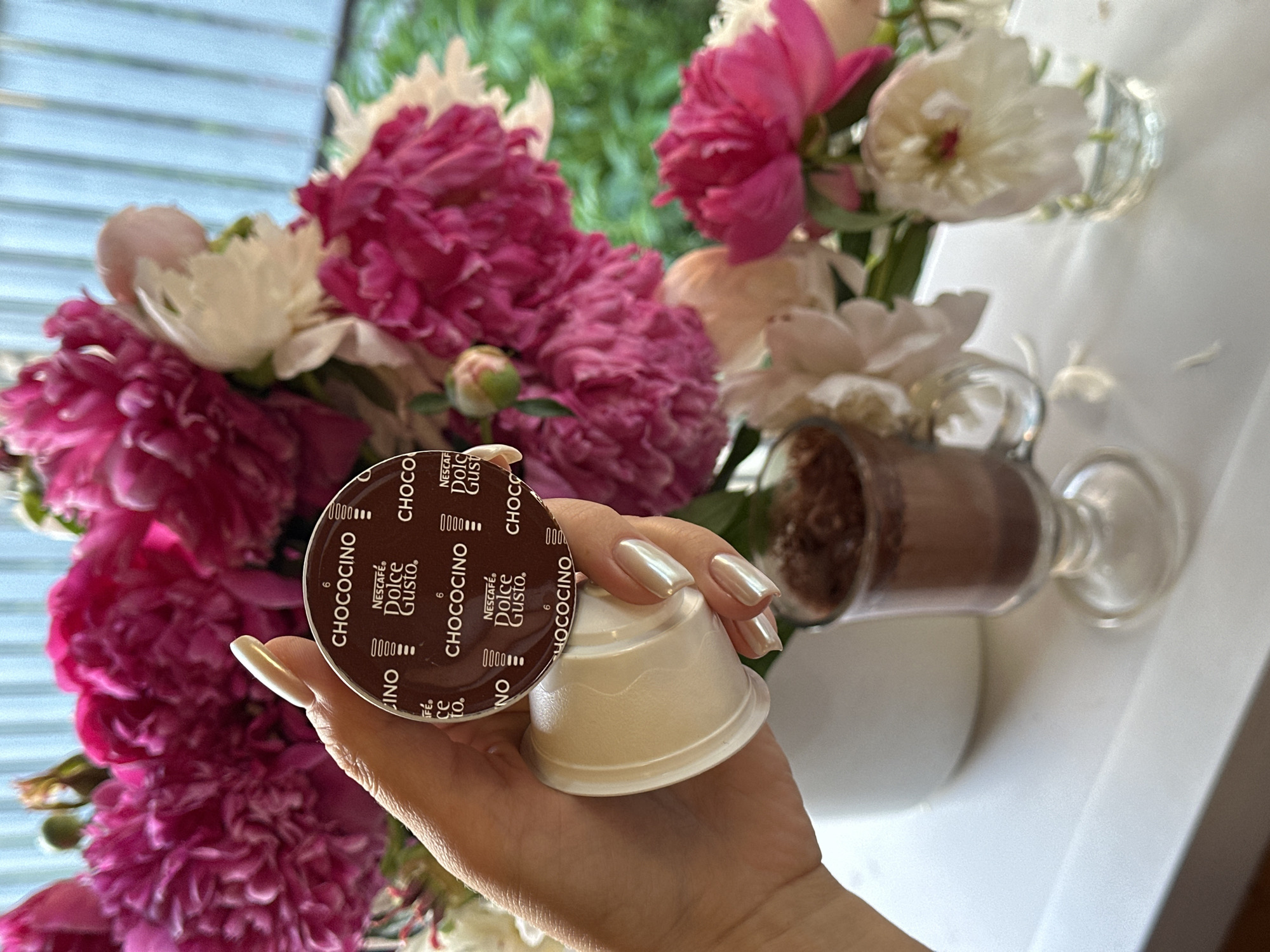 A woman's hand with long, pearlescent manicured nails holds a single serving pod for Nescafé Dolce Gusto Chococcino. The pod is brown with white lettering and a circular pattern. Behind the hand, a bouquet of pink and white peonies is arranged in a white vase. To the right of the vase, a small glass goblet filled with a dark brown liquid, likely hot chocolate, sits on a white surface. The scene appears to be indoors, near a window with a soft blue, slatted blind. The overall impression is a still life with a focus on a beverage and floral arrangement. The image does not provide any information that allows for the localization of the scene in Nizhny Novgorod, Russia.