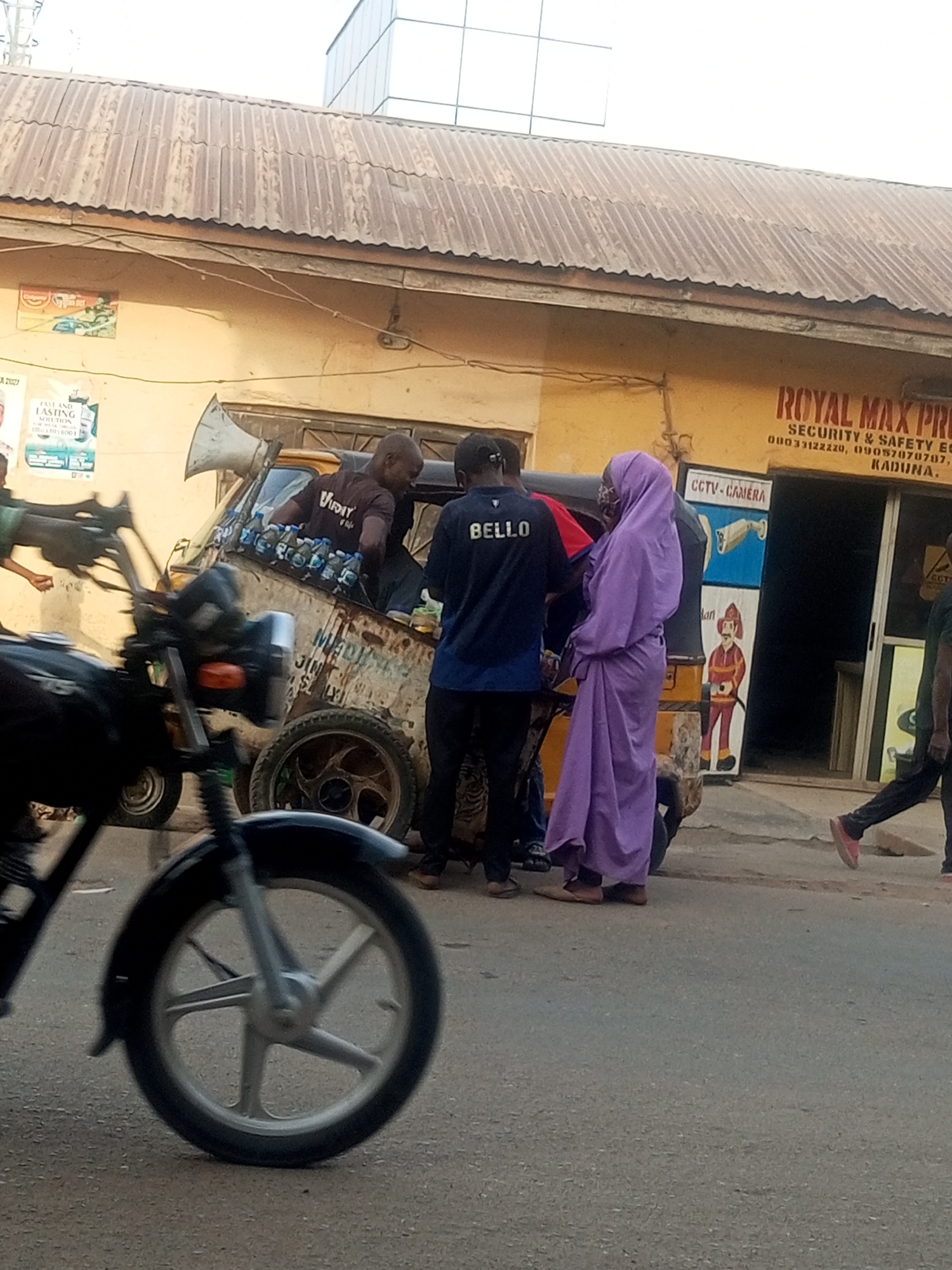 The image depicts a street scene in Kaduna, Nigeria. A three-wheeled motorized vehicle, possibly a "keke napep", serves as a makeshift display for bottled liquids. A man is sitting on top of the three-wheeled vehicle. Another man wearing a dark blue shirt that reads "BELLO" is standing in front of the vehicle. A woman wearing a purple hijab is also present near the vehicle. A motorcycle is partially visible in the foreground, obscuring some of the scene. The backdrop includes a building with signage for "ROYAL MAX PRO SECURITY & SAFETY EC" and a CCTV camera advertisement. A pedestrian is walking past the group.