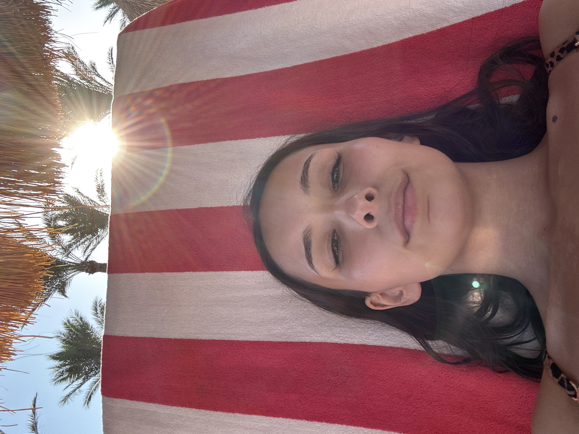A young woman with long dark hair lies on a beach chair covered by a red and white striped towel. She is wearing a leopard print bikini top. The sun shines brightly through palm trees, creating lens flare in the image. The background consists of palm trees and a bright blue sky. The scene appears to be a beach resort in an unknown location in Egypt.