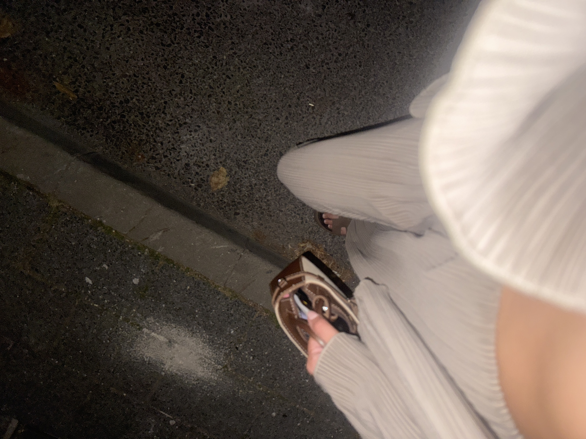 The image shows a person's legs and torso from a high-angle perspective, set against an asphalt ground. The person is wearing a light-colored, pleated outfit and appears to be standing or walking on the ground, which has a rough, textured surface. A small, brown clutch purse with a metallic clasp is held in the person's hand. The purse contains what looks like keys and possibly a tube of lipstick.
The ground surface shows scattered debris, including small leaves and what appears to be a discarded cigarette butt. There is also a white powdery substance scattered on the asphalt, possibly chalk or dust. The scene suggests an outdoor setting, possibly a street or pavement, with a concrete curb visible on the left side. The lighting is dim, indicating it could be evening or nighttime. The overall mood is casual and everyday. No text is visible in the image. The location is Plovdiv, Bulgaria.