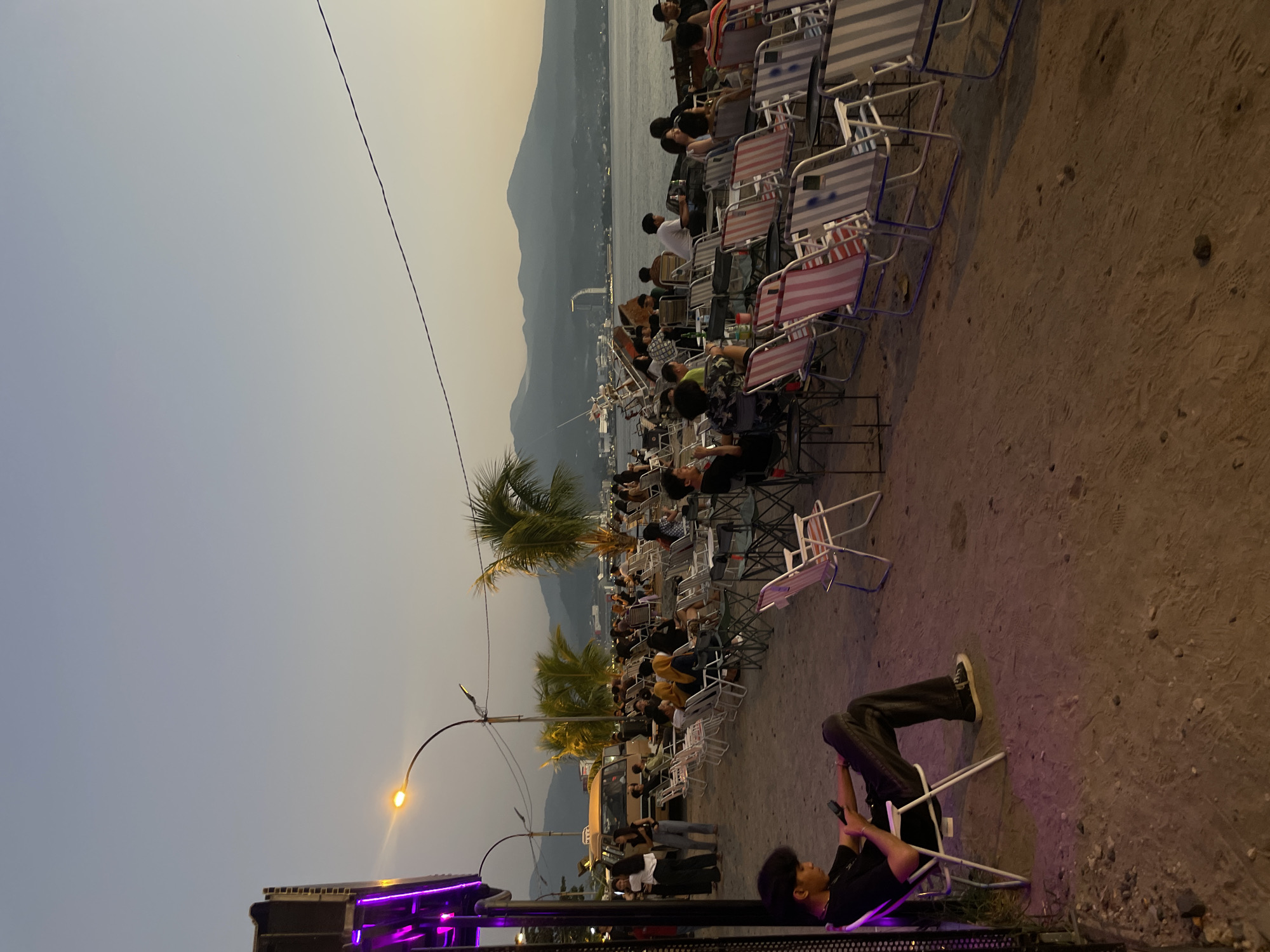 The image captures an outdoor seating area by the sea, likely a cafe or restaurant, with a crowd of people enjoying the evening. The location is identified as Manado, Indonesia, indicated by the presence of palm trees, a distinctive building on the waterfront, and the overall coastal ambiance with mountains in the background.
In the foreground, a young man is seated in a white folding chair, engrossed in his mobile phone. He is wearing a black t-shirt and dark jeans. His posture suggests a relaxed, casual demeanor. The ground is a mixture of sand and gravel.
Further into the scene, rows of folding chairs are arranged, many occupied by patrons. The chairs feature various striped patterns in colors like pink, white, blue, and gray. Some individuals are seen conversing or looking towards the sea. Behind the seating area, a body of water stretches out, with boats and a modern building visible on the opposite shore. The sky is a soft gradient of pale blue and orange, indicative of dusk or early evening.
To the left, a van is parked, and a group of people are standing near it, possibly arriving or departing. A street lamp with a warm yellow light illuminates the area. Purple neon lights are also visible on a structure to the far left, adding a modern, vibrant touch to the scene. Palm trees are scattered throughout the area, contributing to the tropical setting. The mountains in the distance frame the horizon, adding depth to the landscape. The atmosphere appears to be calm and social, with people enjoying a leisurely evening by the water.