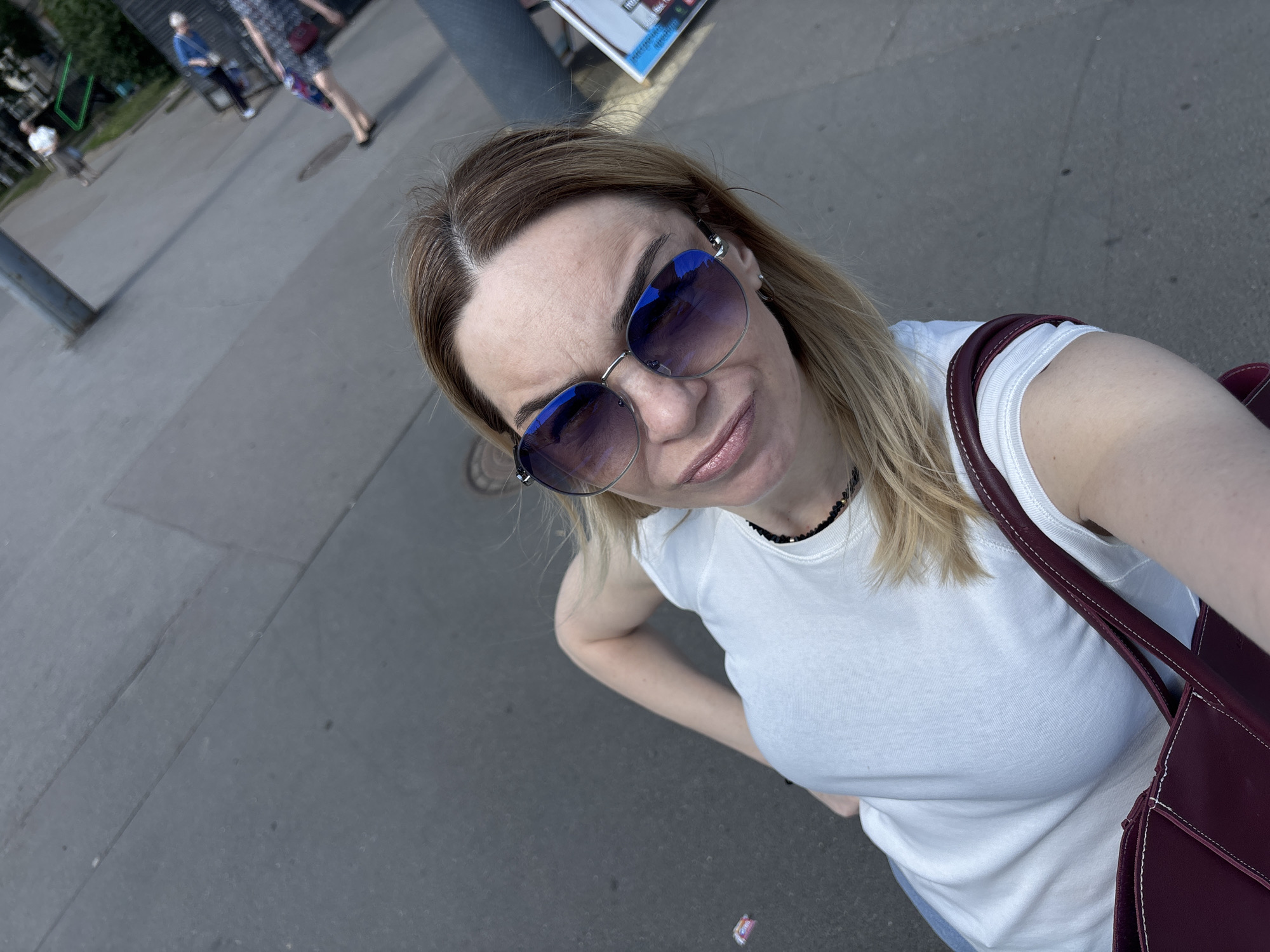This is a high-angle selfie taken by a woman on a sunny day in an urban environment, with the location context provided as Unknown, Russia. The woman, positioned prominently in the foreground, has blonde hair with visible darker roots and is wearing large, blue-tinted reflective sunglasses. She has a subtle smile and her left hand is casually placed on her hip. She is dressed in a plain white crew-neck t-shirt and a dark, possibly beaded, choker necklace. A maroon shoulder bag with contrasting white stitching is visible on her right side, hanging over her shoulder.
The background, largely out of focus, depicts a paved outdoor area, likely a street or sidewalk, indicating a public space. Several blurred figures of people can be seen walking in the mid-ground; one appears to wear a blue top and dark pants, and another a patterned dress. Green foliage and indistinct building structures are visible further behind them. Above the woman's head, a partial view of a sign or poster with illegible text is discernible, suggesting a commercial or informational point. The ground consists of grey asphalt or concrete, with a small piece of colorful litter visible on the path in the lower right corner. The scene conveys a casual, bright daytime outing.