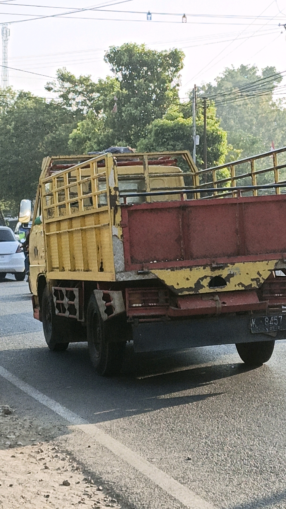 The image shows the rear and side view of a yellow and red flatbed truck on a road. The truck has a cage-like structure on its bed and appears worn, with chipped paint. A license plate with the characters "K 8457" is visible at the back. The truck is in motion on an asphalt road with a white line marking the lane. In the background, there are trees and some overhead wires. A white car is partially visible to the left of the truck. The scene suggests an outdoor environment, likely in an urban or semi-urban area given the presence of roads and other vehicles. The lighting indicates it is daytime, possibly late afternoon due to the angle of the shadows.
