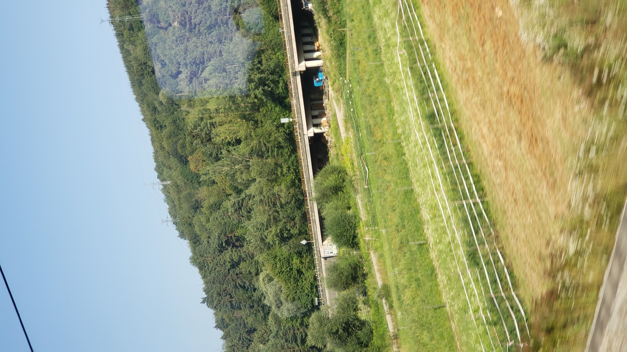 The image captures a rural landscape near Hlboké nad Váhom, Slovakia. A multi-lane overpass, constructed from concrete, dominates the midground, supported by numerous pillars. The overpass is surrounded by a lush green hillside covered in dense forestation. In the foreground, a field of green grass with scattered white wildflowers is visible, bordered by a white electric fence. The sky is clear and blue. Two large electrical towers can be seen on the distant hillside. A blue truck is parked underneath the overpass, and what appears to be construction materials are stacked nearby. The image is taken from a moving vehicle, indicated by motion blur in the foreground.