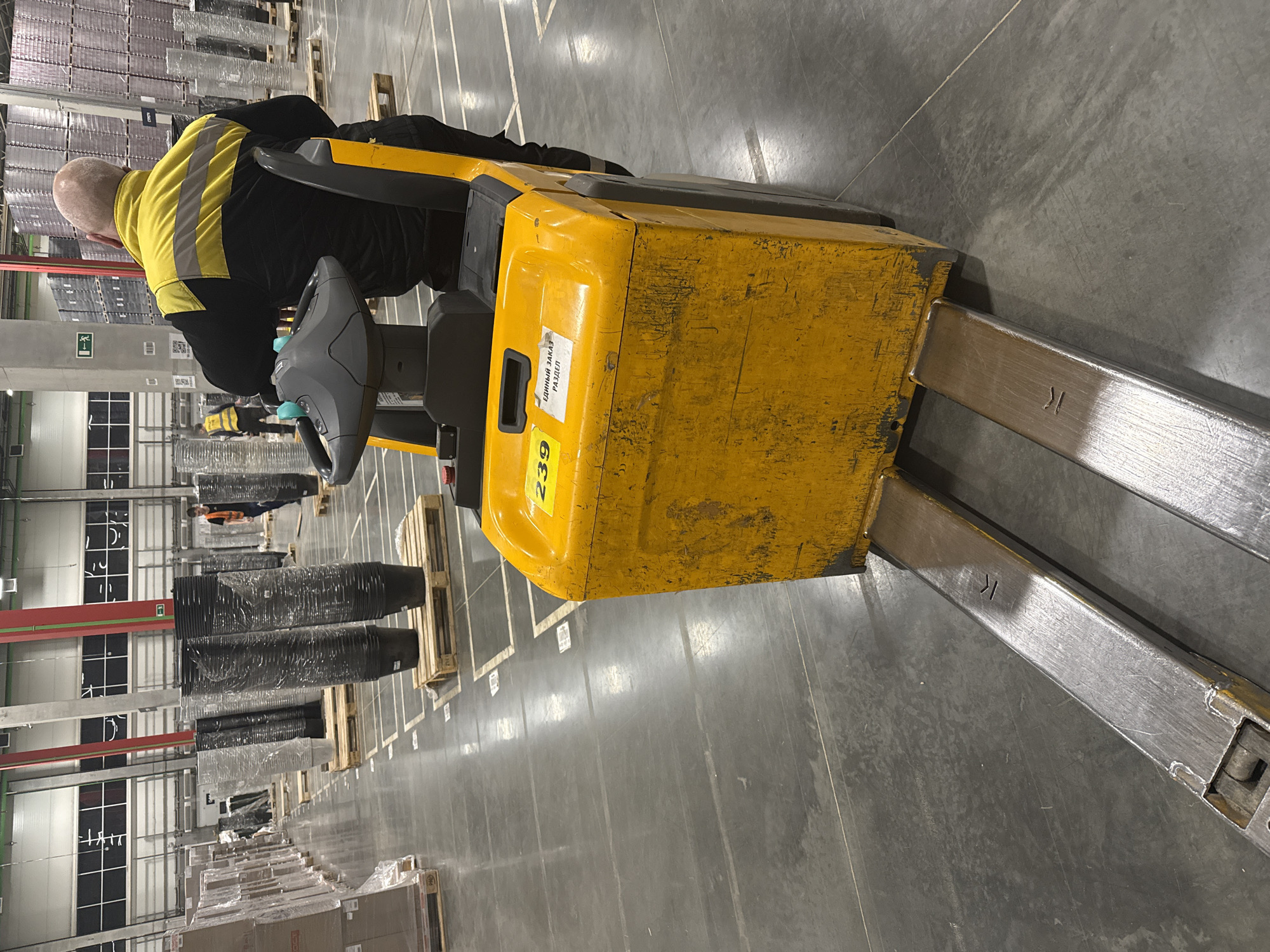 An overhead medium-angle shot depicts the interior of a large warehouse, likely located in Lupanovo, Russia. A bald male, seen from behind and wearing a high-visibility yellow and black jacket, operates a yellow electric pallet jack. The pallet jack is partially numbered with "239" visible on the side. Silver forks protrude from the front of the machine, marked with a "K". Pallets loaded with dark, cylindrical objects wrapped in plastic are visible in the background, along with shelves stocked with goods. White lines are painted on the grey concrete floor. Another individual wearing a high-visibility vest is seen standing in the background.