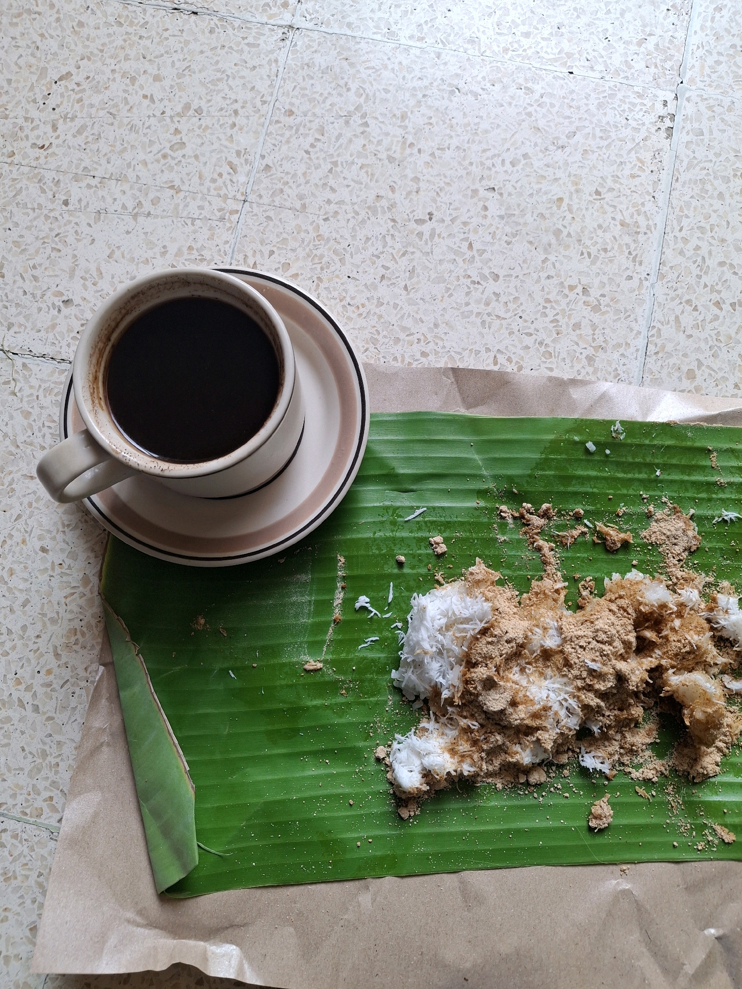 A cup of black coffee rests on a saucer, beside a portion of grated coconut mixed with brown sugar, served on a banana leaf. The banana leaf is placed on a brown paper. The scene is laid out on a light-colored tiled floor. The overall setting suggests a casual breakfast or snack, likely enjoyed outdoors or in a rustic setting, common in Lumajang, Indonesia.