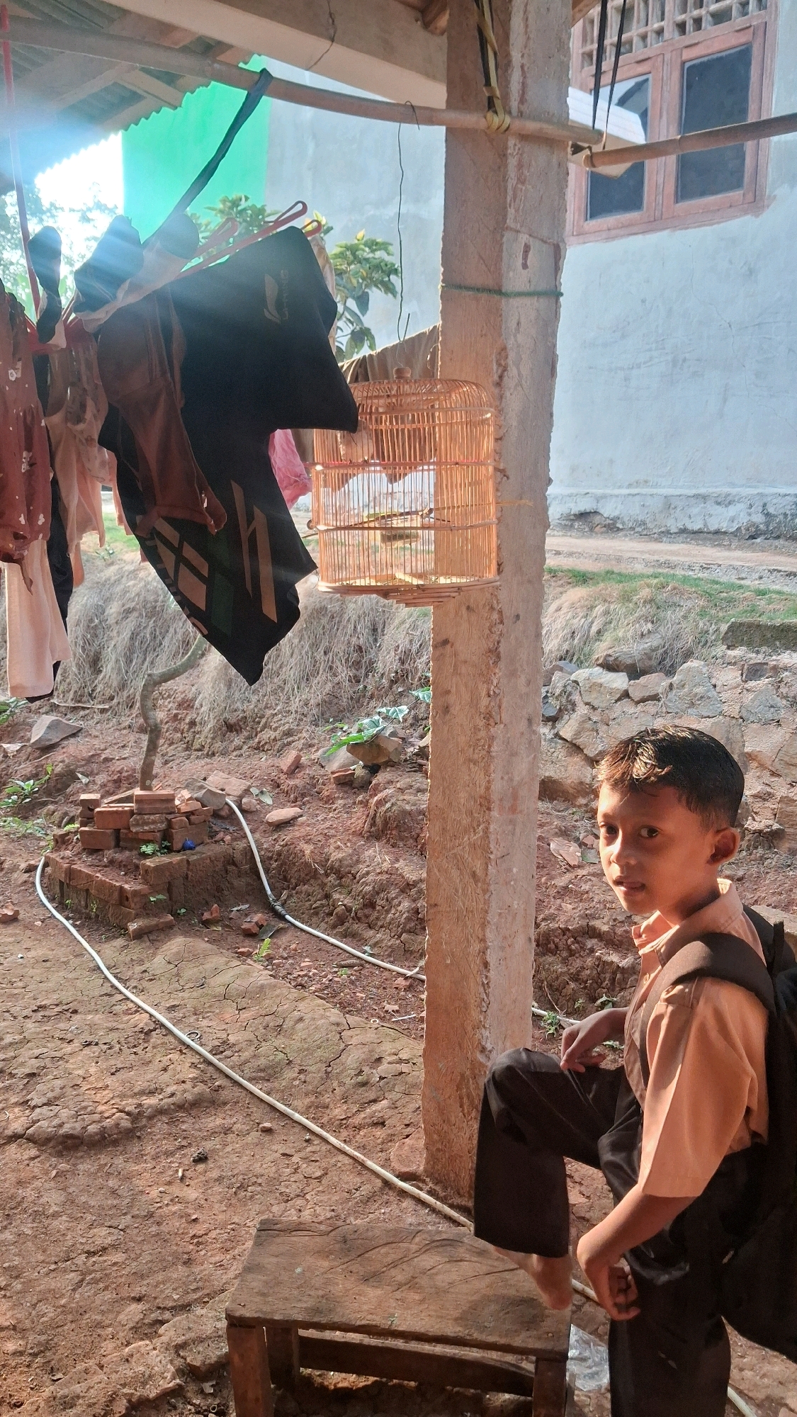 A young boy wearing a peach-colored collared shirt and dark pants sits on a wooden stool in front of a concrete pillar. He is looking directly at the camera with a slight smile. Behind him, laundry hangs from a clothesline under a thatched roof. A small, wooden birdcage is suspended from the pillar, and inside, a bird is visible. The ground is made of red, cracked earth with scattered rocks and some vegetation. In the background, a pale wall with a wooden-framed window can be seen. The scene appears to be in a rural outdoor setting, possibly the yard of a house in Karang Sari, Indonesia.