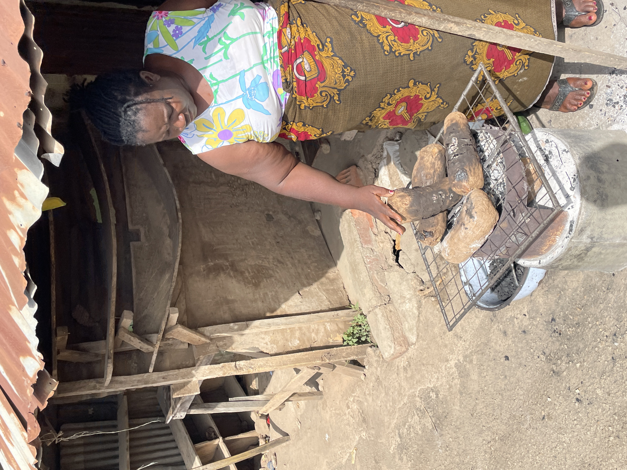 A woman is captured from an overhead perspective, cooking root vegetables on a charcoal grill outdoors or in a semi-enclosed space. She is wearing a white floral-patterned top and a dark wrapper with red and gold patterns. Her dark hair is styled in braids, and her gaze is directed towards the food.
Her right hand is extended, seemingly adjusting or checking one of the large, irregularly shaped, dark-skinned root vegetables (likely yams) resting on a metal grill. Below the grill is a simple, bucket-like charcoal stove, which contains visible embers. The ground is a mix of concrete and dirt.
In the background, to the left, there are rough wooden planks forming a utilitarian structure, possibly shelving or support beams. Corrugated metal roofing or siding is visible on the far left. Bright sunlight illuminates the scene, suggesting it is daytime. The woman wears black sandals with red painted toenails, and the word "GUCCI" is faintly visible on the strap of one sandal. A small cluster of green weeds grows near the base of one of the wooden structures. The overall setting implies a practical, informal cooking area, consistent with the provided location context of Jos, Nigeria.