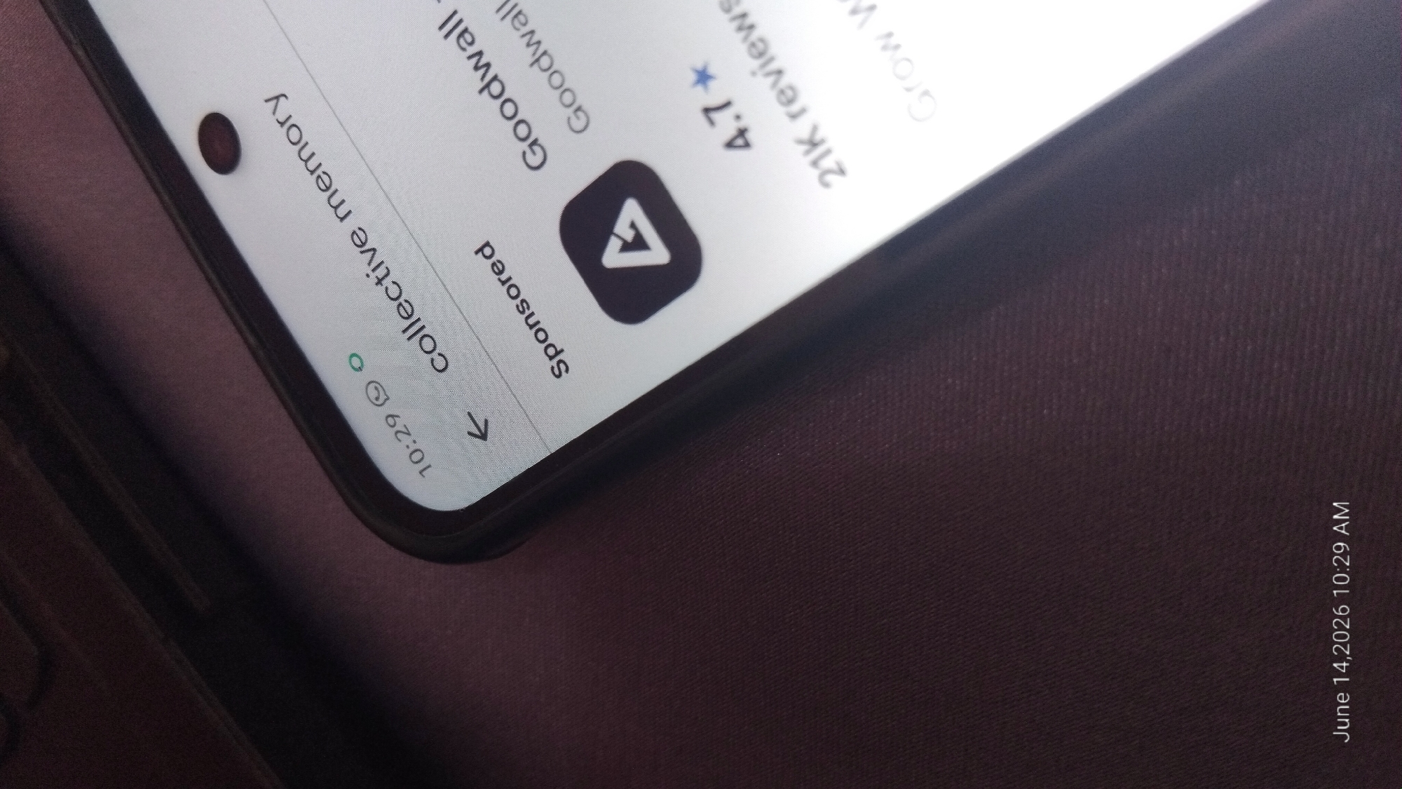 A close-up, angled shot shows a smartphone screen displaying a Google Play Store listing. The screen shows a time of "10:29" and the text "collective memory." Below this, "Sponsored" is visible, followed by a black icon with a white triangle inside. The app name appears to be "Goodwall" and the rating is "4.7*" with "21K reviews." The bottom left corner of the image shows a timestamp: "June 14, 2026 10:29 AM." The smartphone is resting on a dark, textured surface, possibly fabric or a desk. Part of a dark object, likely a laptop, is visible in the upper left corner.