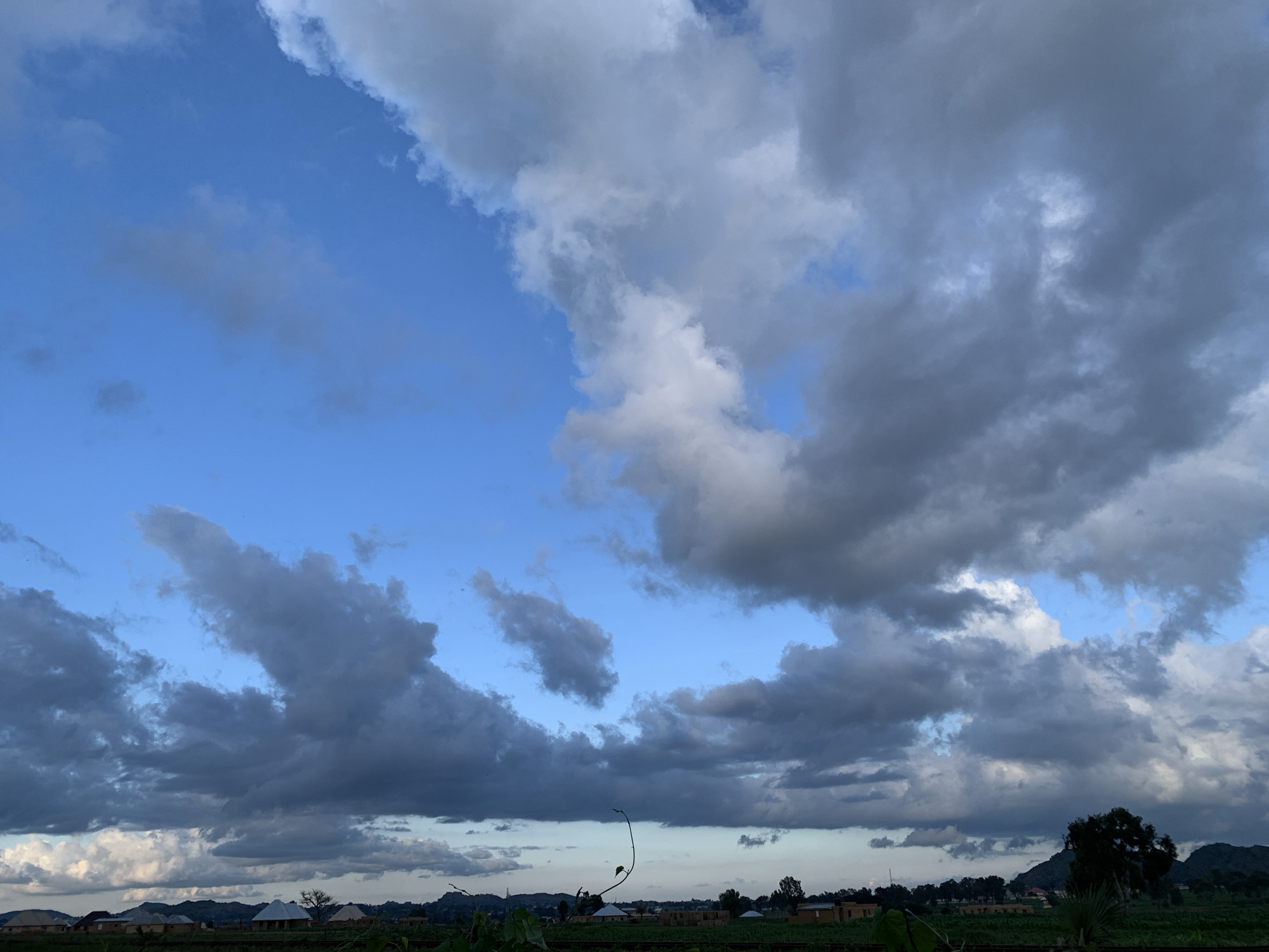 This is a photograph taken in Kuru Karama, Nigeria, featuring a dramatic sky filled with a mix of blue sky and dark, heavy clouds. The clouds dominate the upper half of the frame, with some appearing light and fluffy while others are dense and grey, suggesting an approaching storm or recent rainfall. Below the sky, a landscape unfolds with a hint of human settlement. Small, traditional buildings with peaked roofs are visible along a low horizon, interspersed with trees and lush greenery. The overall scene evokes a sense of natural beauty and the vastness of the sky over a rural Nigerian setting.