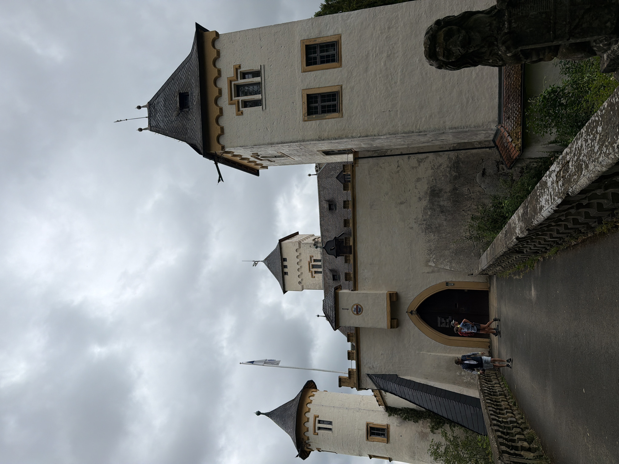 A castle stands on a hill against a cloudy sky. The castle is white with a yellow trim and dark grey roofs. It has several towers, including one on the right with a tall, pointed roof and two smaller towers on the left. A drawbridge leads to the main entrance, which is an arched doorway. Two people are walking across the drawbridge, one in a blue shirt and shorts, and another in a red shirt and dark shorts. A third person in a grey vest and shorts is walking away from the camera on the drawbridge. A stone lion statue stands to the right of the drawbridge. The castle appears to be well-maintained and is surrounded by trees and greenery. The overall impression is one of history and grandeur.