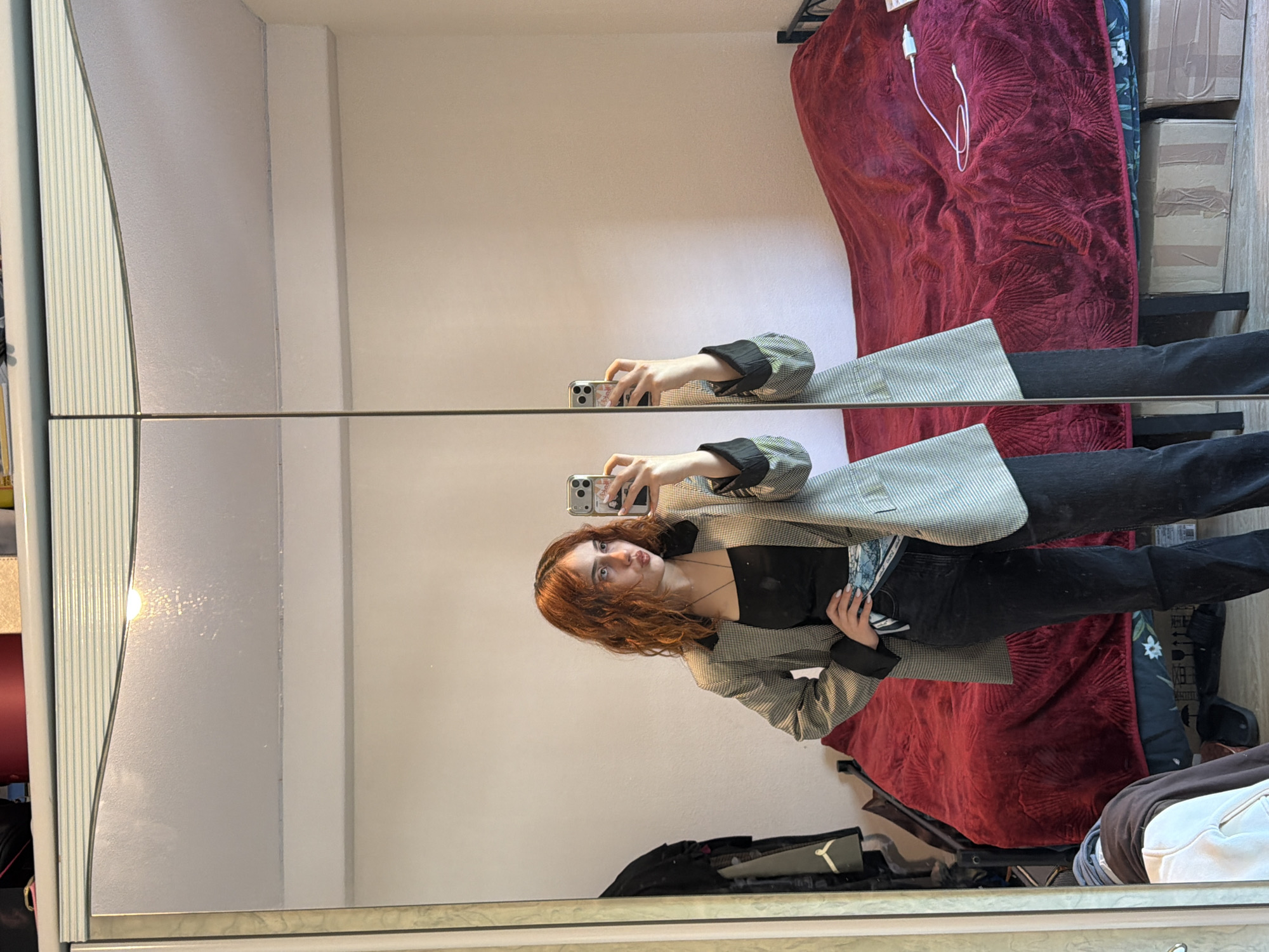 An indoor mirror selfie captures a young person with long, reddish-brown wavy hair, looking directly at the camera with a playful, slightly puckered expression. The individual is holding an iPhone with a patterned case to take the picture. They are dressed in a black strapless top, dark-colored jeans, and a light-colored blazer with a subtle houndstooth or plaid pattern and dark cuffs. Light-colored nail polish is visible on their fingers.
The selfie is taken in front of a large mirror, possibly part of a wardrobe, which has a vertical division and a horizontal dividing line at the person's hip level. The room has light-colored, possibly off-white, walls. In the background, reflected in the mirror, is a bed covered with a dark red, richly textured blanket or duvet, on which a white charging cable is resting. To the right of the bed, stacked cardboard boxes are visible, suggesting a storage or cluttered area. The overall setting appears to be a bedroom or personal space. The location context is Strasbourg, France, although no specific visual cues confirm this in the image itself. There is no clearly discernible text visible in the image.