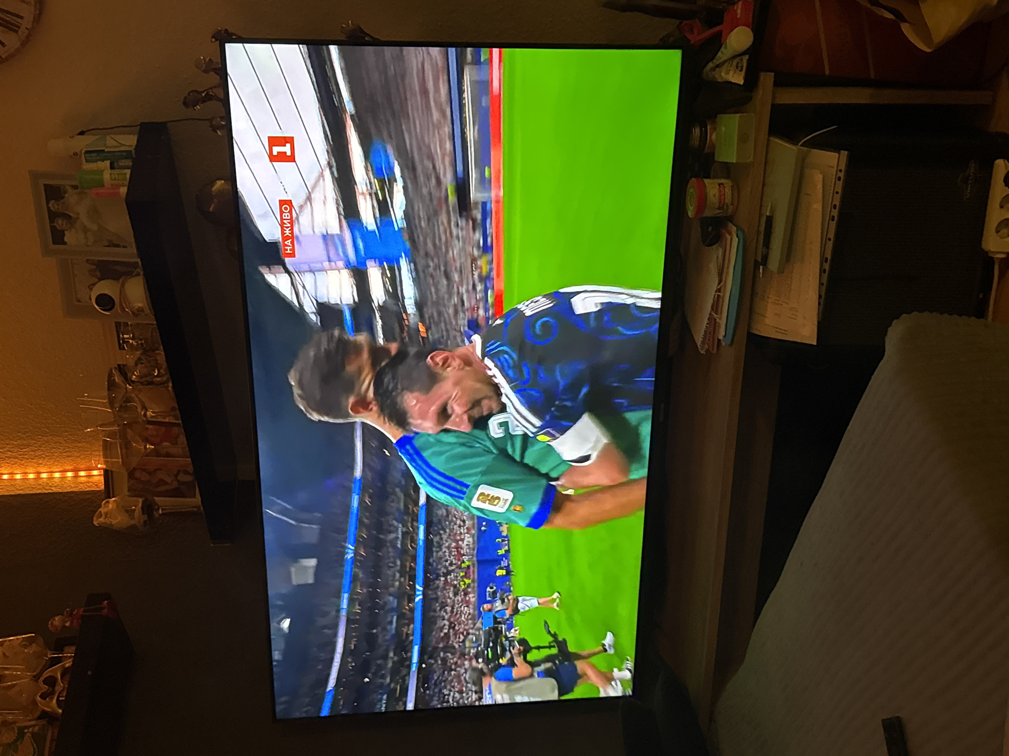 The image shows a television screen displaying a soccer match. Two male soccer players are prominently featured, with one wearing a green jersey and the other a blue and white striped jersey. The player in the blue and white jersey appears to be Lionel Messi, based on his distinctive features. The match is being broadcast live, indicated by the "НА ЖИВО" (On Air) text and the number "1" displayed on the screen, suggesting a major sporting event. The television is placed on a wooden table, which also holds various items such as papers, a jar, a green box, and other miscellaneous objects. In the background, above the television, shelves are visible with decorative items and picture frames. The overall setting suggests a domestic environment where the soccer match is being watched. The image was captured in Lübeck, Germany.