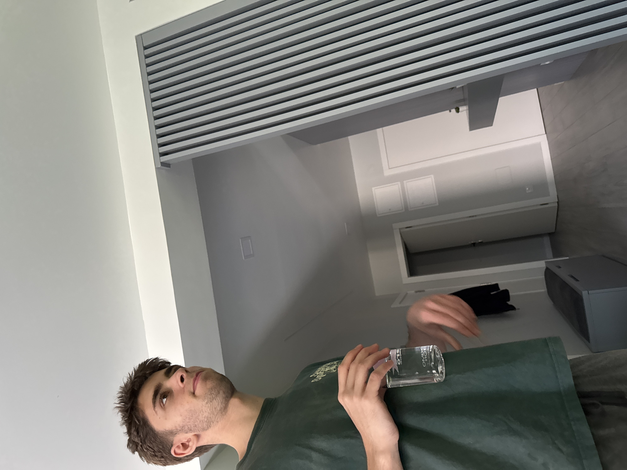 A man stands in a modern interior, possibly in Mirandela, Portugal. He holds a glass vial of clear liquid in his hands. In the background, there is a white door and a gray device on the floor. The ceiling is composed of horizontal gray slats.