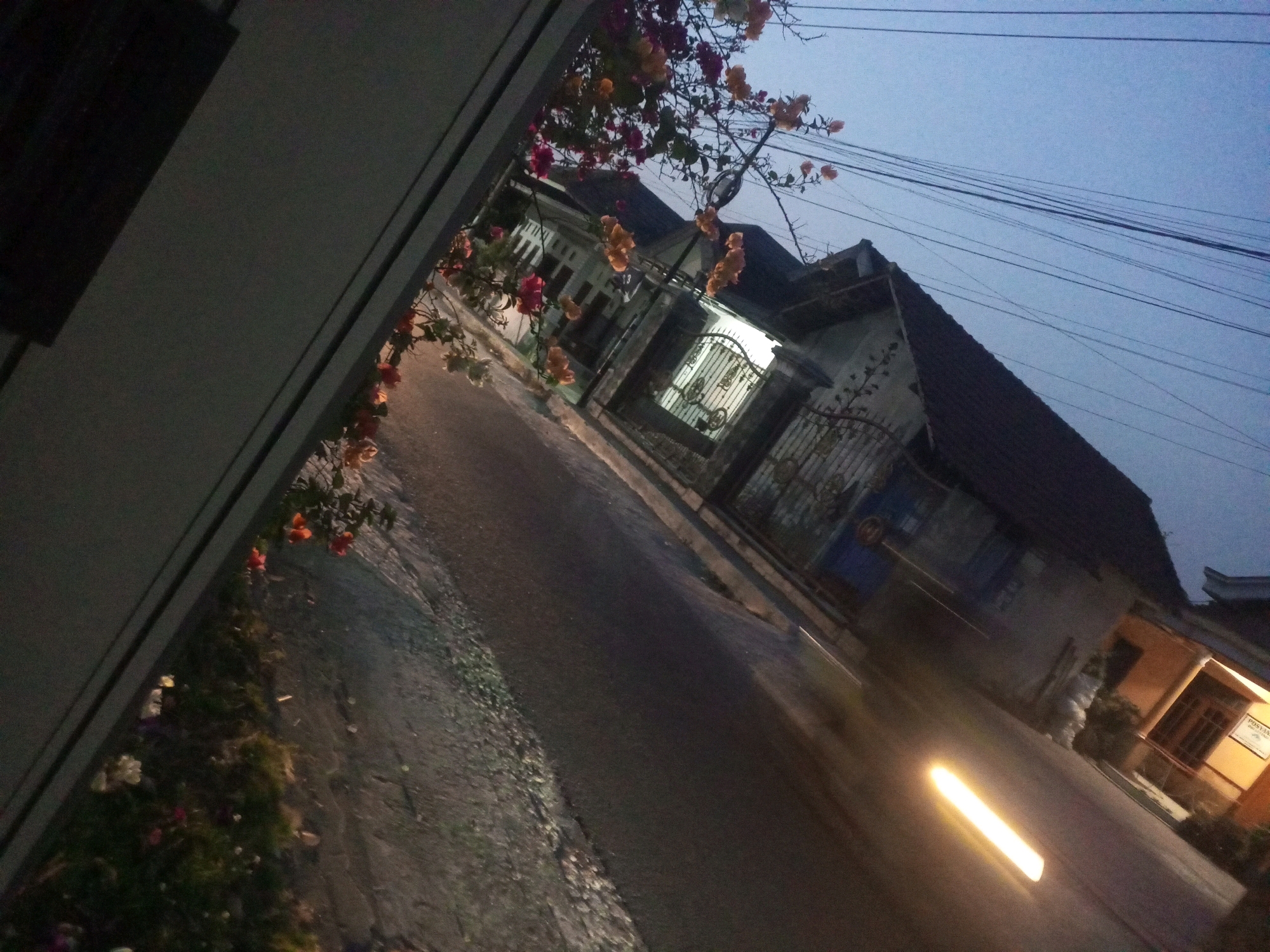The image depicts a street scene at dusk or dawn in Ngasem, Indonesia. A narrow asphalt road runs diagonally from the foreground towards the background. On the left side of the road, a white building wall is visible, angled towards the viewer. Above the wall, flowering plants with red, pink, and orange blossoms are hanging over the scene. Across the street, several houses with dark roofs are present, one featuring a brightly lit gateway with an ornate metal fence. Electrical wires are strung across the sky above the houses. A streaky light, possibly from a moving vehicle's headlights or a streetlamp, streaks across the road in the mid-ground. The sky is a pale blue, indicating low light conditions. The overall impression is of a quiet residential street at the transition between day and night.