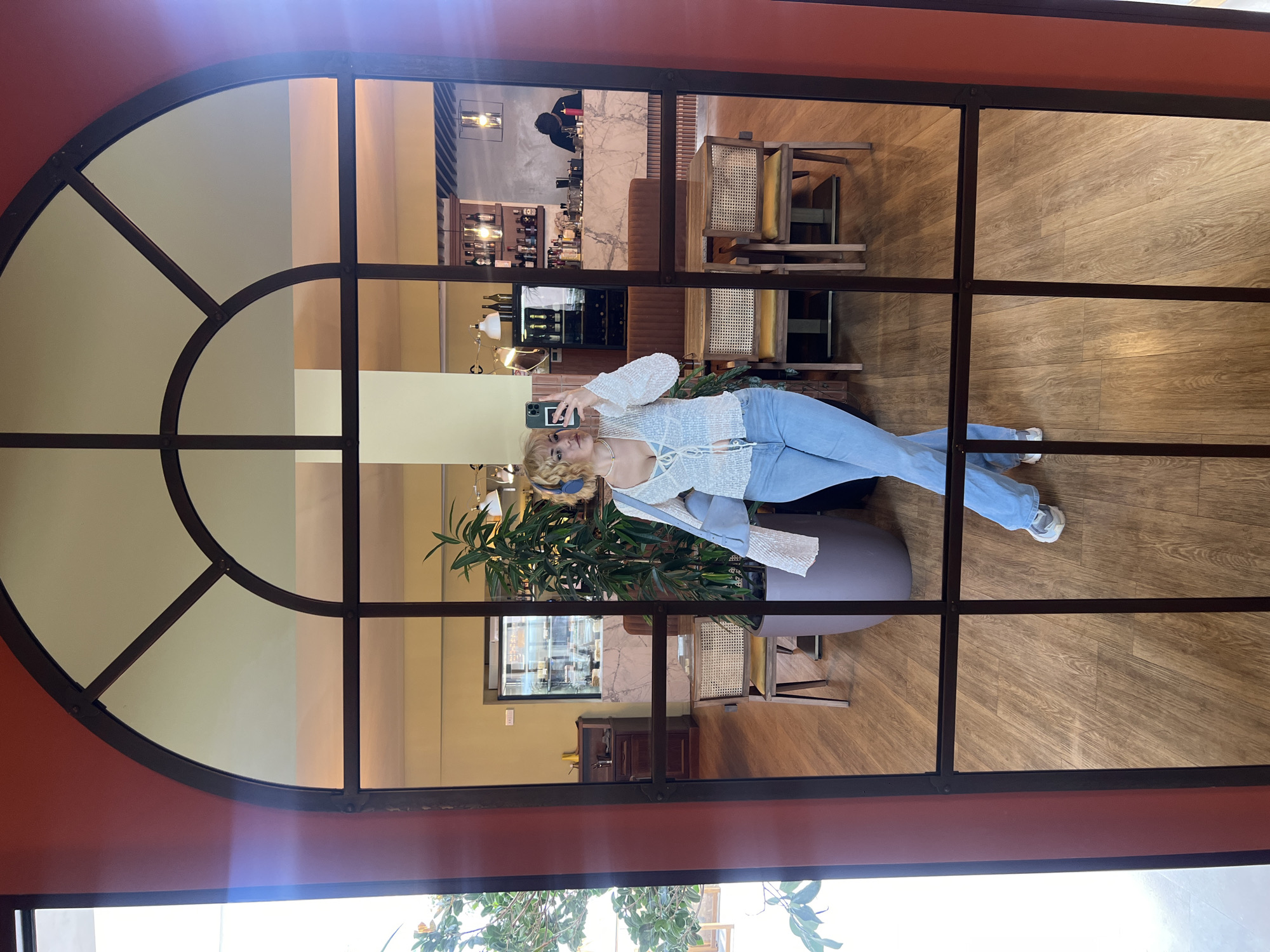 A woman stands in the reflection of a large, arched, multi-paned window within a building in Boreti, Montenegro. The woman is holding a phone, possibly taking a selfie, and is dressed in a light-colored, lacy top and light blue jeans. The interior of the building is visible through the window, revealing a bar area, tables and chairs, and other interior design elements. Outside the window, trees with green foliage are visible. The window frame is dark, and the surrounding wall is painted in earth tones.