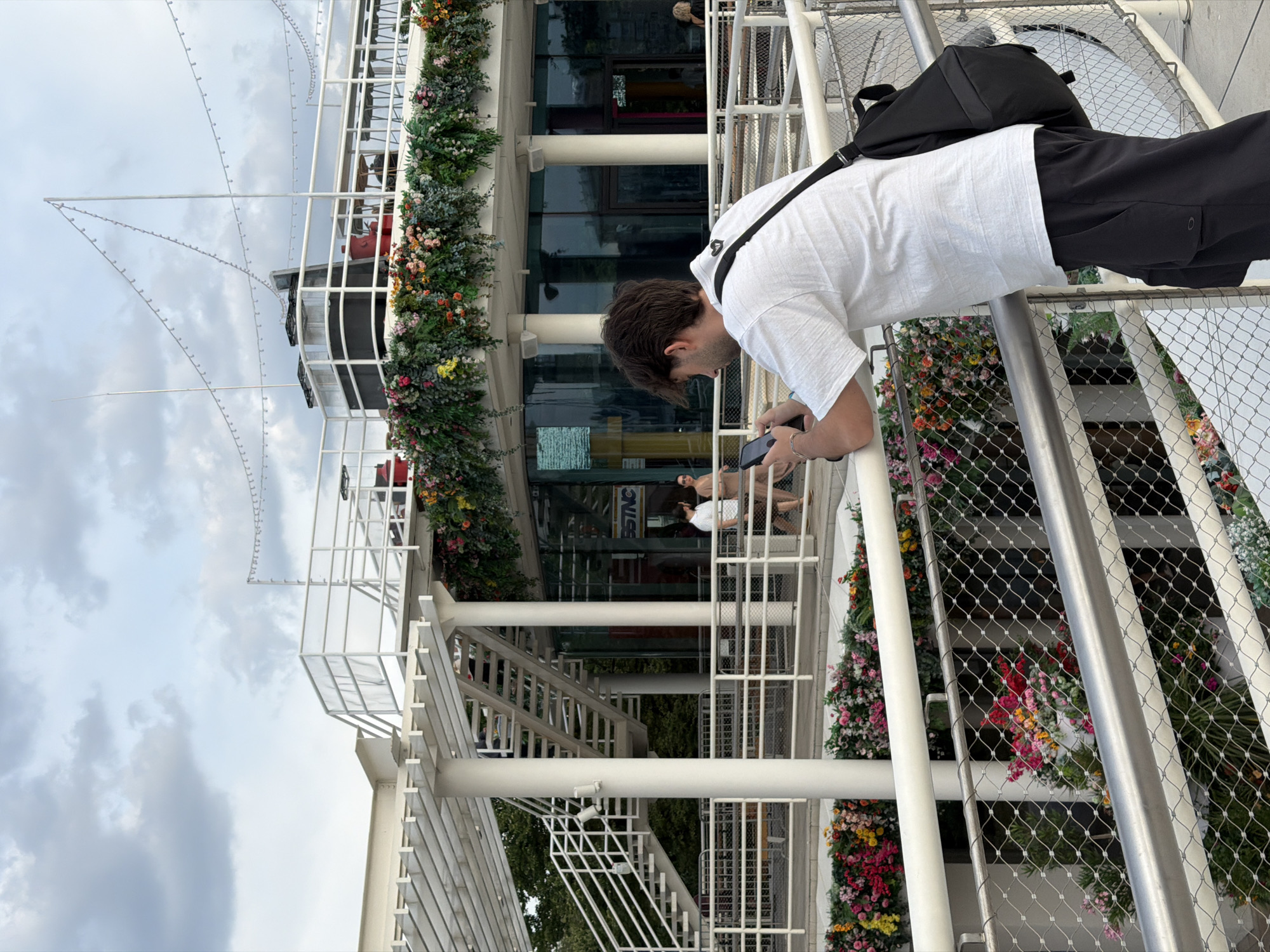 The image shows a man looking at his phone while leaning on a railing. He is wearing a white t-shirt and black shorts, with a black bag slung over his shoulder. Behind him, a building with a glass facade is decorated with abundant floral arrangements. The building appears to be a restaurant or event space. In the background, other people are visible walking and sitting. The sky is cloudy. The location is the Gorky Park in Moscow, Russia.