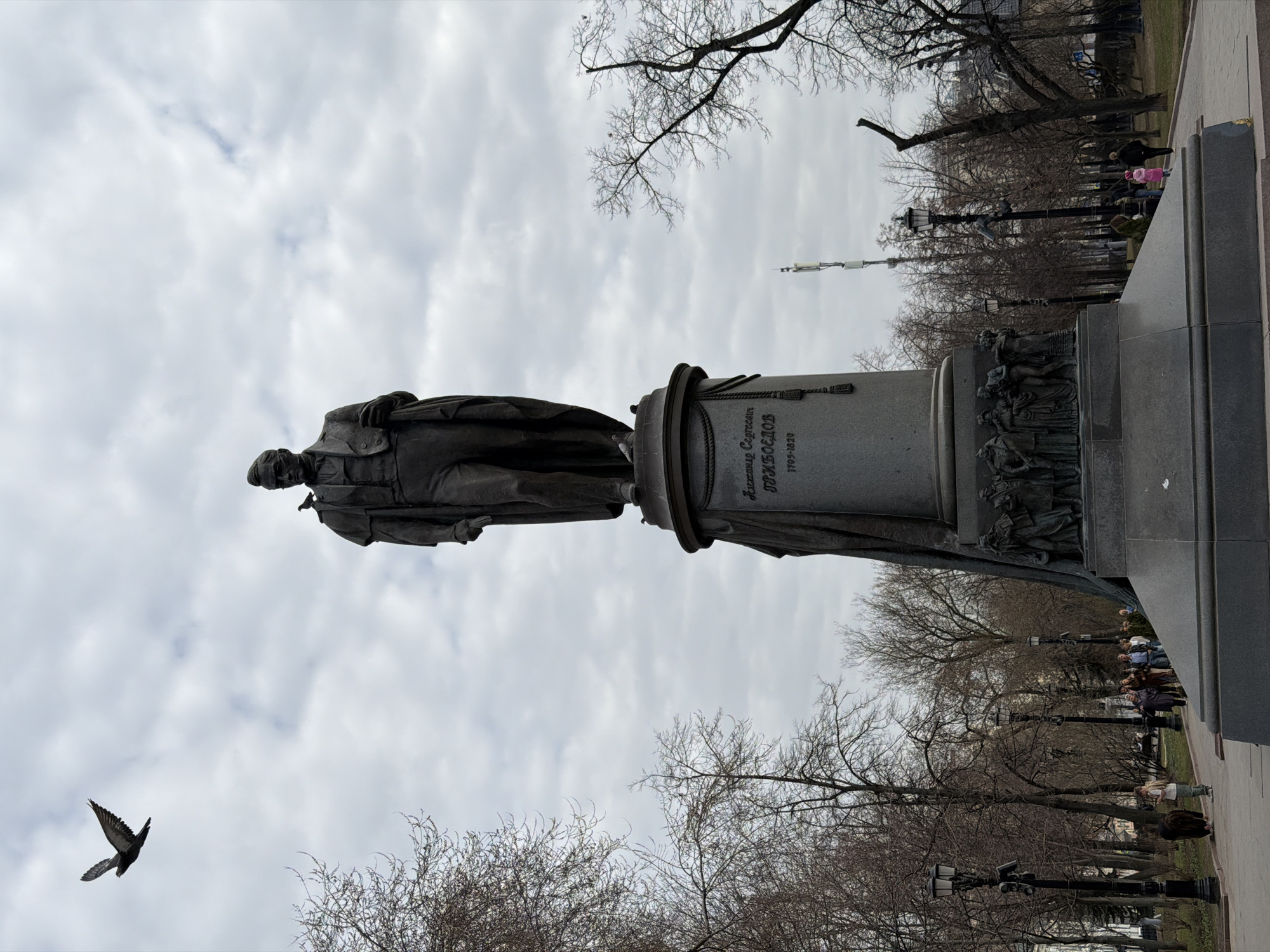 The image depicts a statue in a park on an overcast day. The statue is of Alexander Griboyedov. The statue is mounted on a pedestal with inscriptions in Russian and a sculptural frieze at the base. People are walking through the park in the background. A bird is in flight in the foreground. This statue is located in Moscow, Russia.