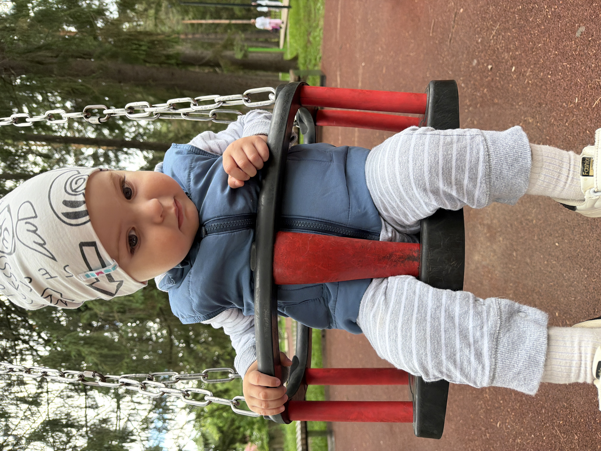 An infant sits on a playground swing in Votkinsk, Russia. The child is wearing a light-colored beanie with black drawings, a blue puffer vest over a striped long-sleeved shirt, and grey striped pants with white socks. The swing seat is black with red supports. The ground beneath the swing is covered in reddish-brown mulch. In the background, trees and other playground equipment are visible. Two adults are walking in the distance.