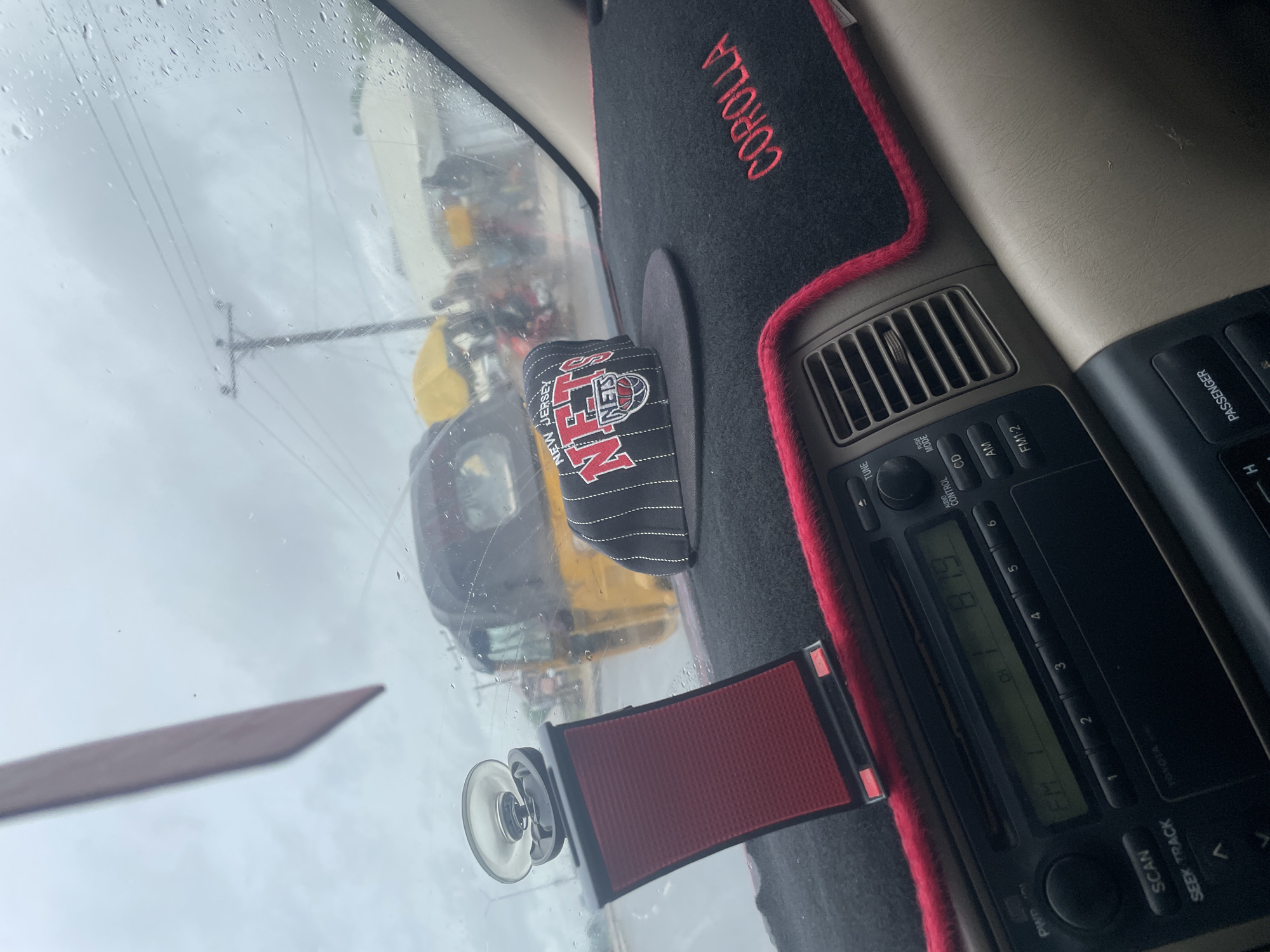 The image depicts the interior of a vehicle, looking through the windshield on a rainy day. A black dashboard mat with a red trim covers the dashboard, and a New Jersey Nets baseball cap rests on it. A red phone holder with a suction cup is attached to the windshield. The radio is visible with the station displayed as "FM 1 87.9". Outside, a yellow auto-rickshaw, also known as a "Keke Napep," is driving on a wet road. Several people and market stalls are visible in the background. The scene suggests a bustling environment in Jalingo, Nigeria, possibly during a market day or a busy thoroughfare. The overall atmosphere is one of everyday life in a Nigerian city, characterized by traffic, street vendors, and the pervasive presence of auto-rickshaws.