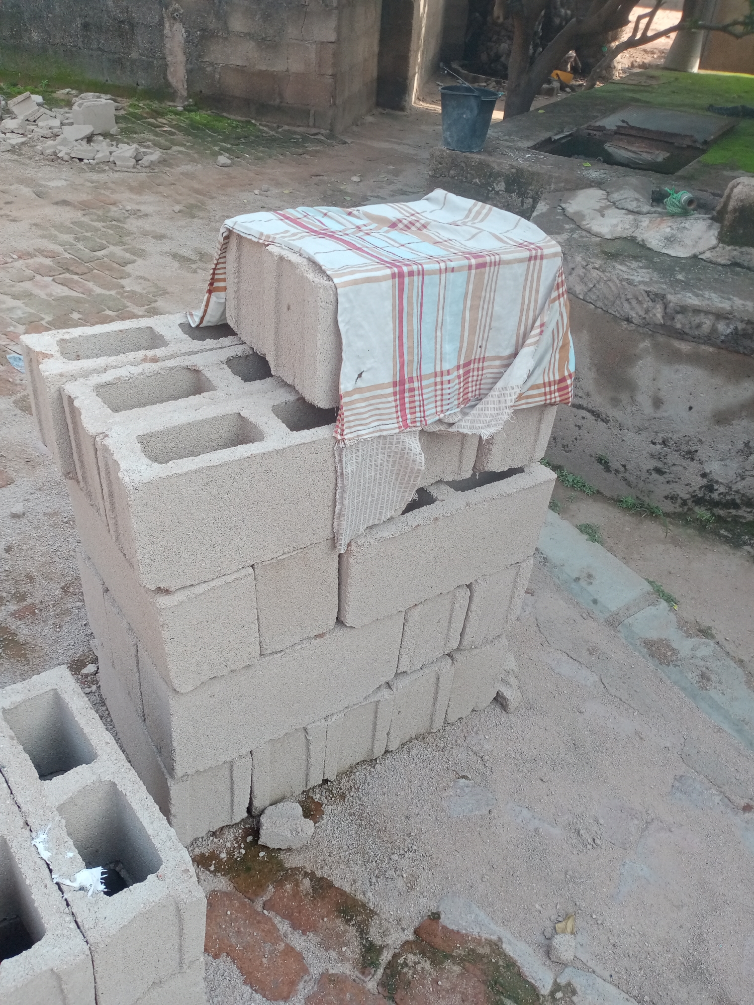 The image depicts an outdoor scene in Jalingo, Nigeria. In the foreground, a stack of light grey concrete blocks is arranged on a dirt and sand surface. A checkered cloth, featuring red, white, and brown stripes, is draped over the top of the stack of blocks. To the left of the main stack, another concrete block is placed horizontally. The ground is composed of dirt, sand, and some red bricks. In the background, a weathered grey stone wall forms the backdrop of the scene. There are various other elements including scattered debris, a dark grey bucket, and some vegetation. The overall impression is of a construction or storage area.