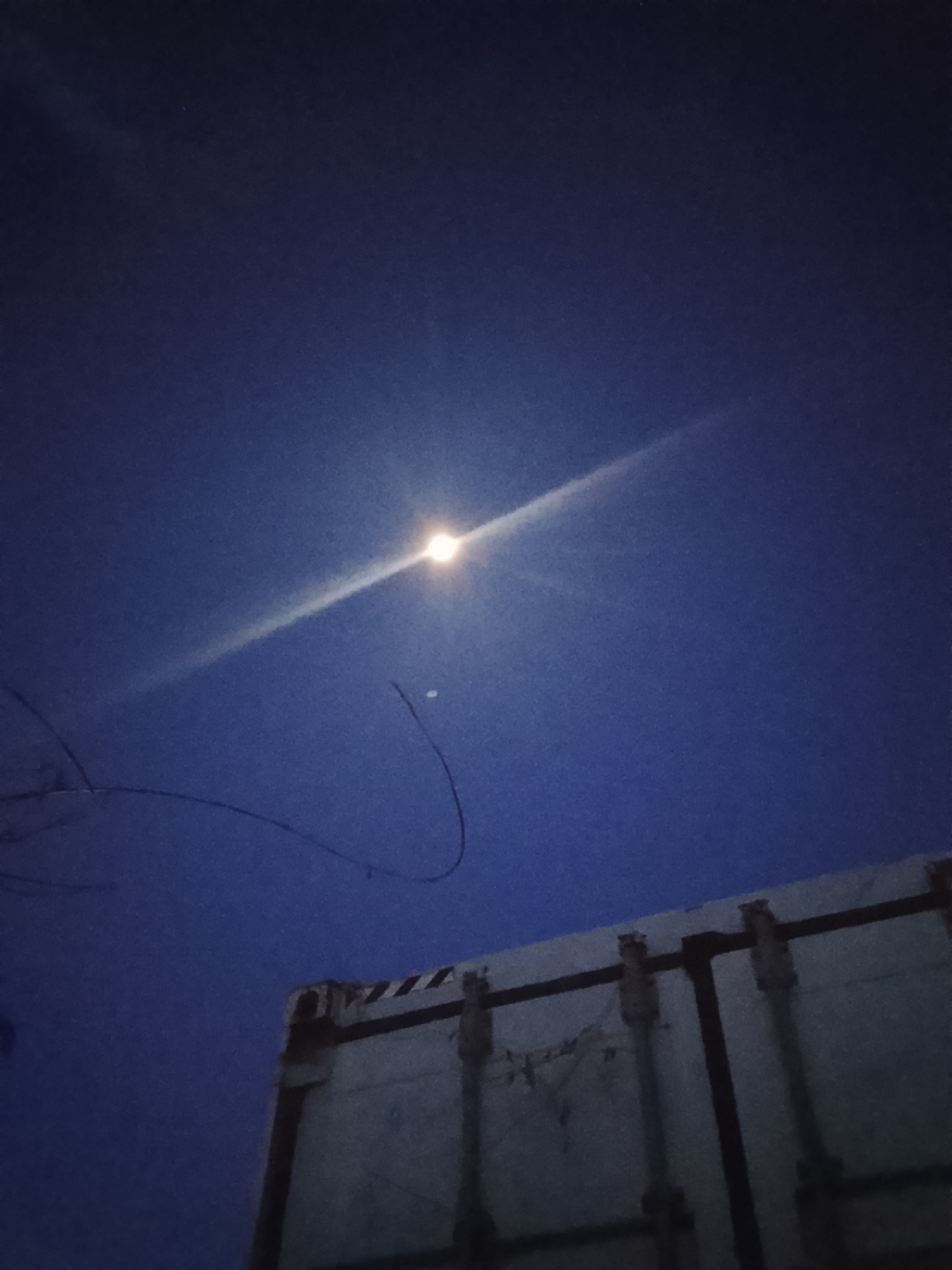 The image shows a night scene in Altagracia, Venezuela. The sky is dark blue with a bright light source, possibly the moon, emitting a radiant beam. A faint object is visible near the moon. In the foreground, the upper corner of a shipping container is visible on the right side. It is light-colored with dark metal fittings and a striped pattern at the top. On the left, thin, dark, curved lines, likely tree branches or wires, stretch across part of the sky.