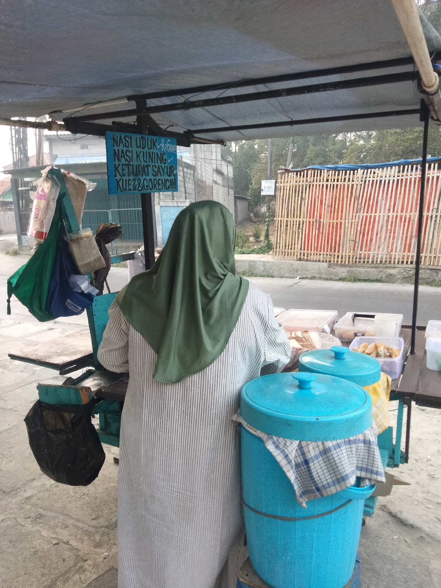 A street vendor, wearing a green headscarf and a striped dress, stands behind a food cart in Pisangan Jaya, Indonesia. The cart is covered with a tarp and displays a sign advertising "NASI UDUK RABU KUNING KETUPAT SAYUR KUE & GORENGAN." The vendor is preparing food, with various containers and items visible on the cart. A black garbage bag hangs from the side of the cart. In the background, a bamboo fence and trees can be seen, along with some buildings. The scene depicts a typical street food stall in a residential or commercial area of the city.