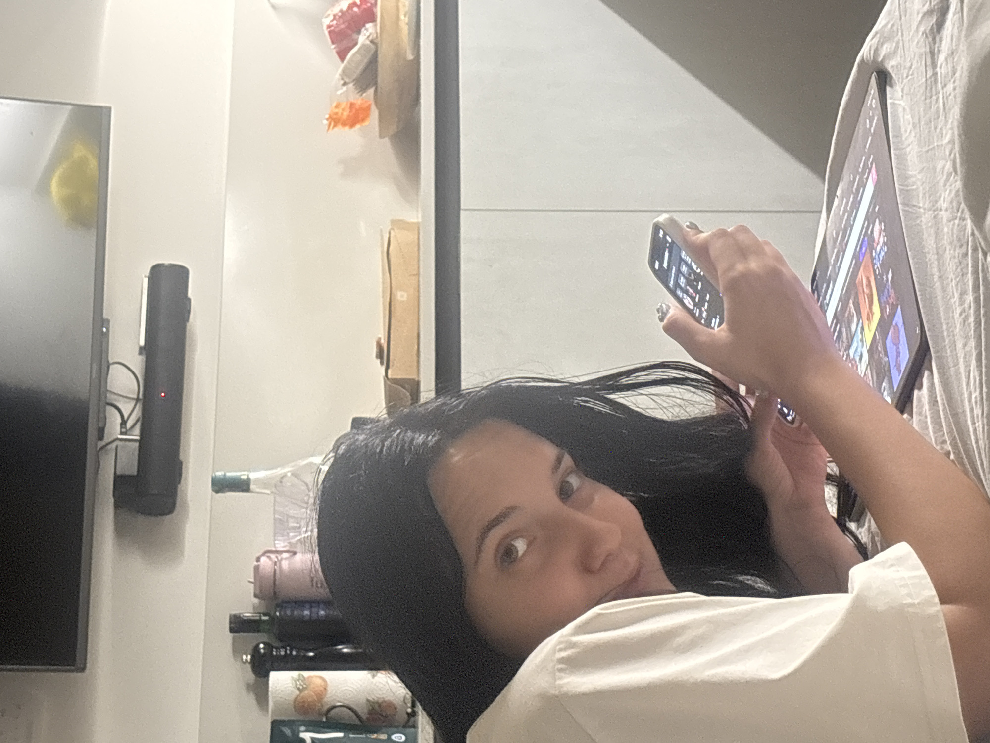 The image shows a young woman with long black hair lying on a bed, looking at her phone and a tablet. She is wearing a white t-shirt. In the background, there are kitchen cabinets, shelves with bottles and containers, and a television mounted on the wall. The lighting suggests it is indoors, possibly in a residential setting. The location is not specified within the image itself, nor is there any information that would place it in Cupra Marittima, Italy.