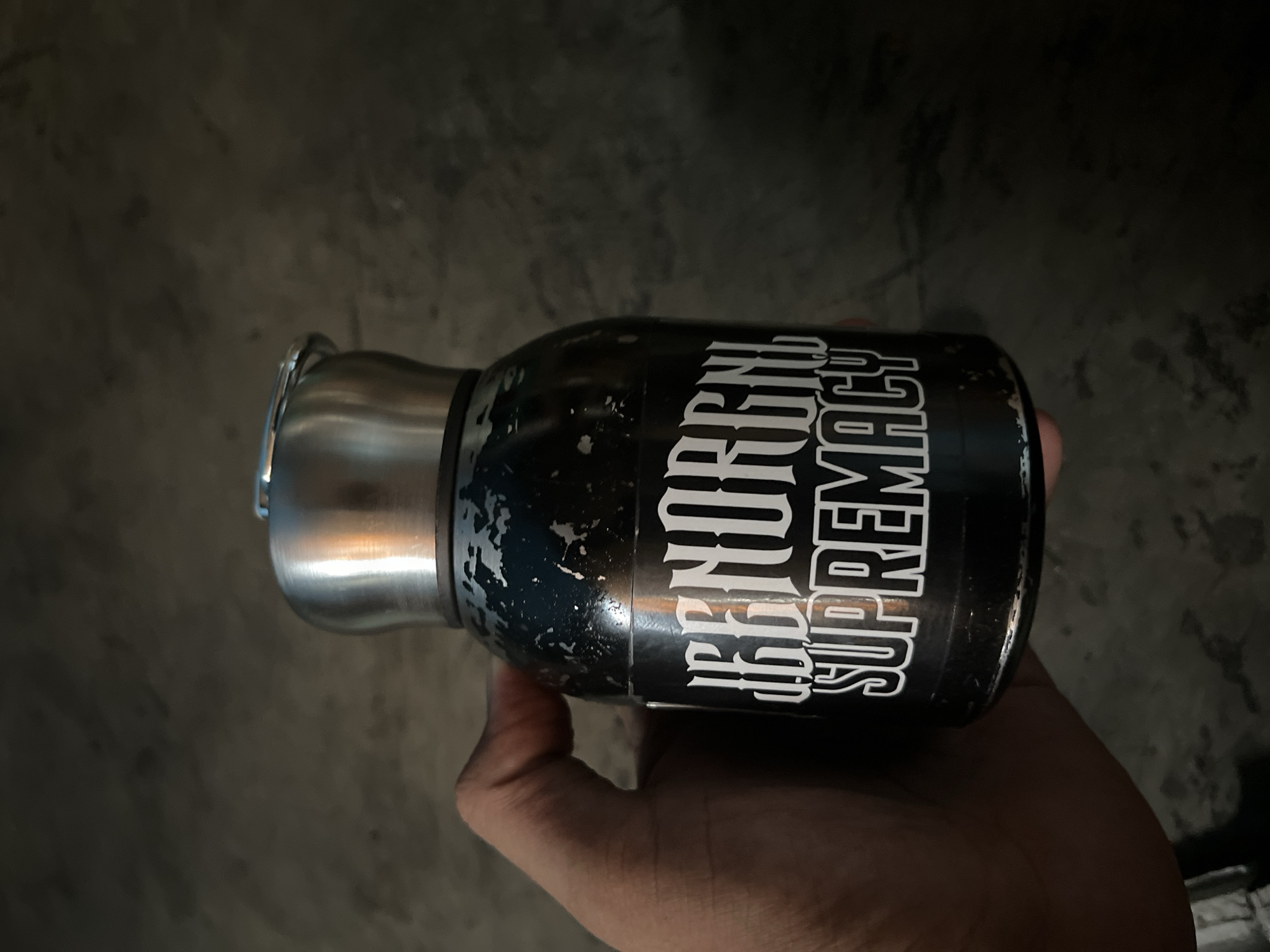 A hand is holding a black thermos with a metallic lid. The thermos has white lettering on it that says "IRREGULAR SUPREMACY". The background is a dark, textured surface. The image was taken in Central Jakarta, Indonesia.