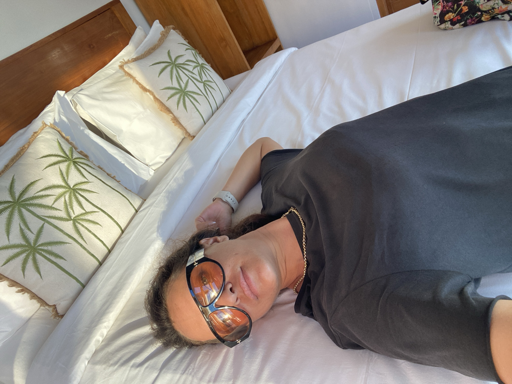 A woman is lying on a bed, facing upwards with her head tilted to the side. She is wearing a dark t-shirt and sunglasses with orange lenses. A gold chain is visible around her neck. Her left wrist has a smartwatch. The bed is covered in white sheets, and there are several white pillows with a palm tree design. The headboard of the bed is made of wood. The scene appears to be indoors, possibly in a bedroom. The lighting suggests it is daytime, with sunlight illuminating the bed and pillows. The overall mood seems relaxed and peaceful. The location is identified as Nusa Penida, Indonesia.