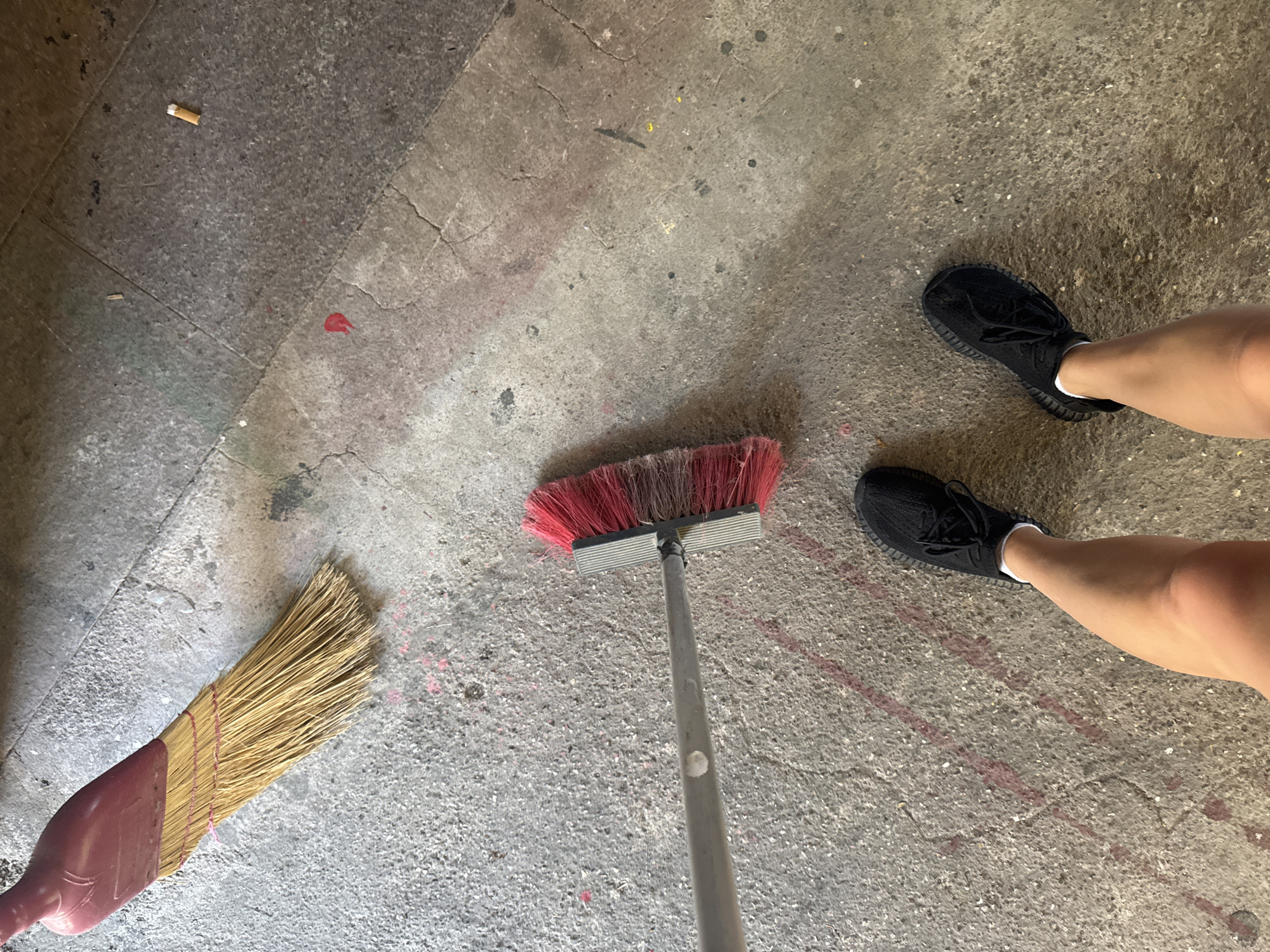 A first-person perspective shot shows a person's legs and feet, wearing black athletic shoes, standing on a rough, grey concrete surface. The ground is marked with scattered red spray paint or chalk lines, and there are also a few smudges of pink and yellow. To the left, a broom with a red plastic handle and natural bristles lies on the ground. Further to the left and slightly out of focus, another broom with a red head and natural bristles is also visible. In the upper right corner, a single cigarette butt rests on the concrete. The overall environment suggests an outdoor urban setting, possibly a street or courtyard. The image does not provide any specific visual information to pinpoint the location as Aksu, Turkey.