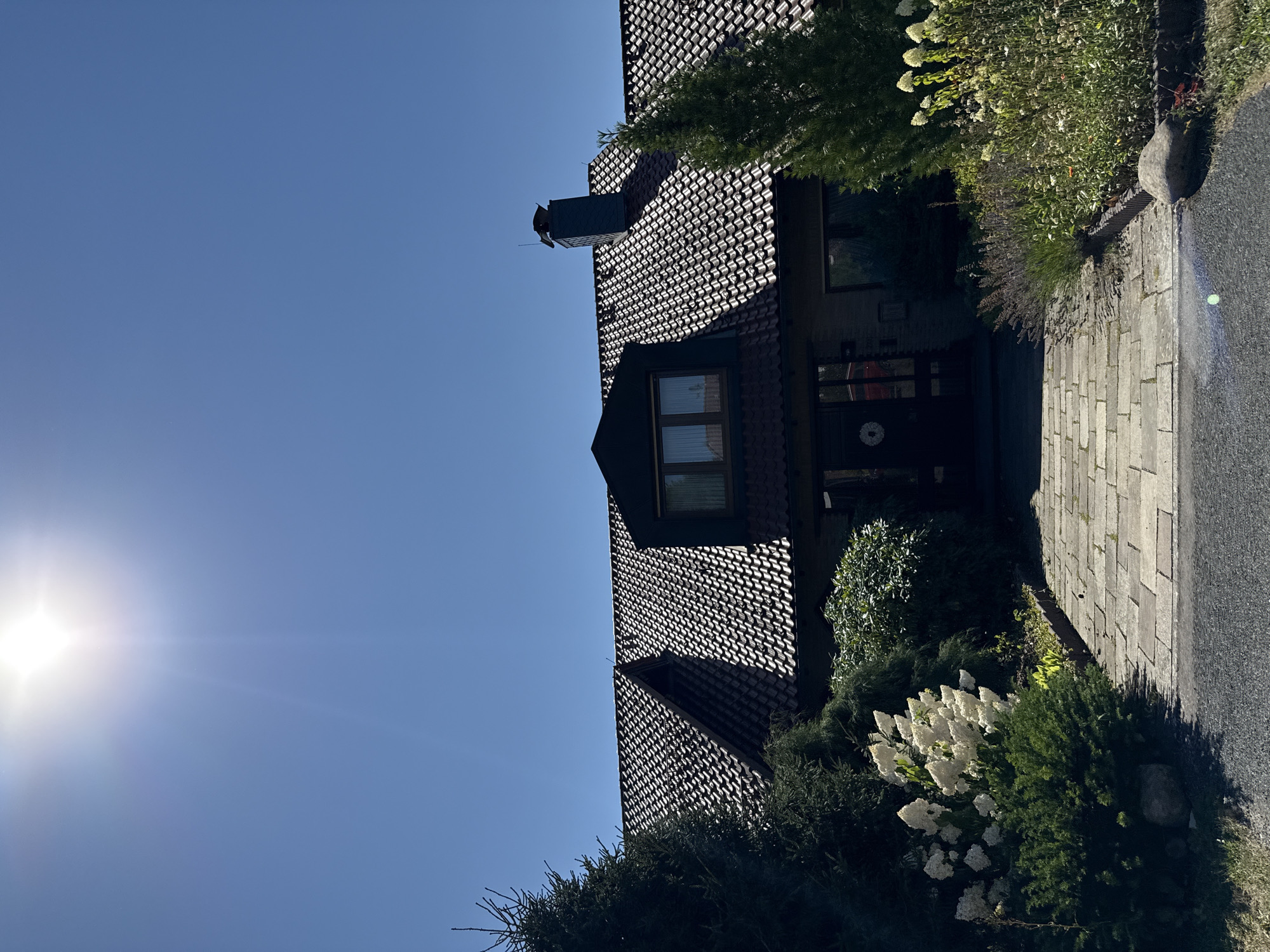 The image shows a sunny day in front of a house located in Bad Lauterberg im Harz, Germany. A paved walkway leads up to the front entrance of the house, which has a tiled roof and a prominent chimney. The house is surrounded by greenery, including conifer trees and flowering bushes. The sun is shining brightly in the clear blue sky, casting shadows on the roof of the house and the walkway. The overall scene depicts a serene residential setting.