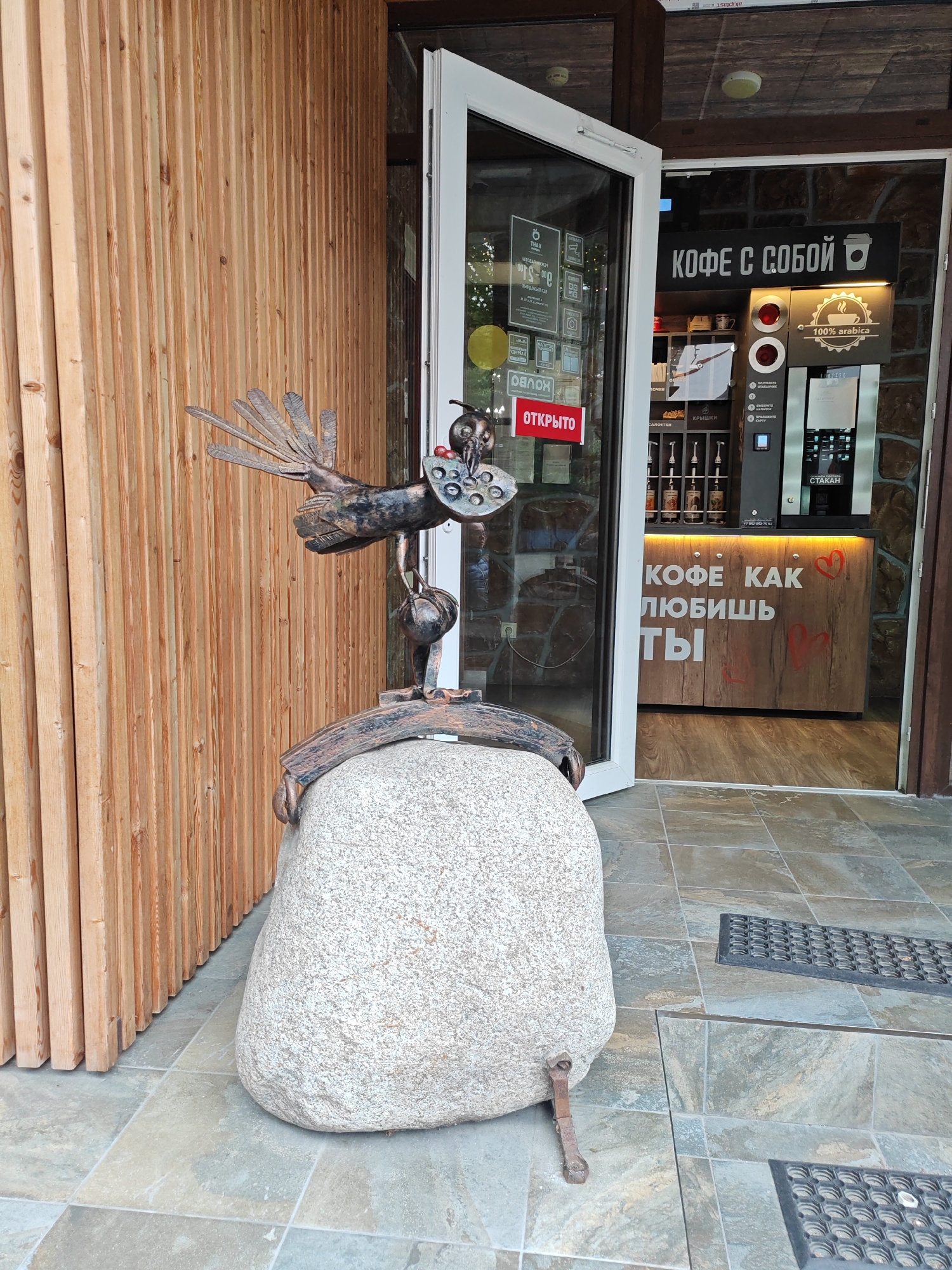 A metal sculpture of a bird perched on a large boulder sits outside a coffee shop. The shop has a wooden facade and an open glass door leading inside. The bird sculpture is detailed, appearing to be made of dark metal with outstretched wings and a small sphere held in its beak. The boulder is rough and grey. Inside the coffee shop, a counter displays coffee-making equipment, cups, and various syrups. A sign above the counter reads "КОФЕ С СОБОЙ" (Coffee To Go) with a coffee cup icon. Another sign on the counter says "100% arabica". A red sign on the glass door reads "ОТКРЫТО" (Open). The coffee shop's interior walls are made of stone and wood. The floor outside the shop is tiled with large, square grey and beige tiles, with black matting in some areas. The overall scene suggests a pleasant, possibly daytime setting, with the coffee shop appearing to be open for business. The artwork outside adds a unique, whimsical touch to the establishment.