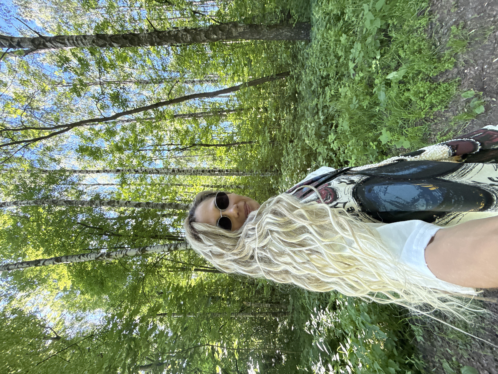 This image captures a single adult female in a dense forest or wooded area under natural daylight. The individual is positioned in the foreground, looking over their right shoulder towards the camera, with their right arm visible holding the camera for a selfie. They have long, wavy blonde hair extending down their back and over their right shoulder. The person wears dark, round sunglasses, a white short-sleeved t-shirt, and an outer garment featuring a large, intricate design in dark blue, black, white, and red, with some shimmering elements. The background is dominated by numerous trees with green foliage, including several birch trees with distinct white and dark bark. Patches of clear blue sky are visible through the tree canopy, indicating a sunny day. The ground is covered with dense green undergrowth, including various plants and grasses, with a visible dirt path in the lower right portion of the frame. The lighting is bright and natural, suggesting a clear day in a natural, wooded environment. The location is a forest in Unknown, Unknown.
