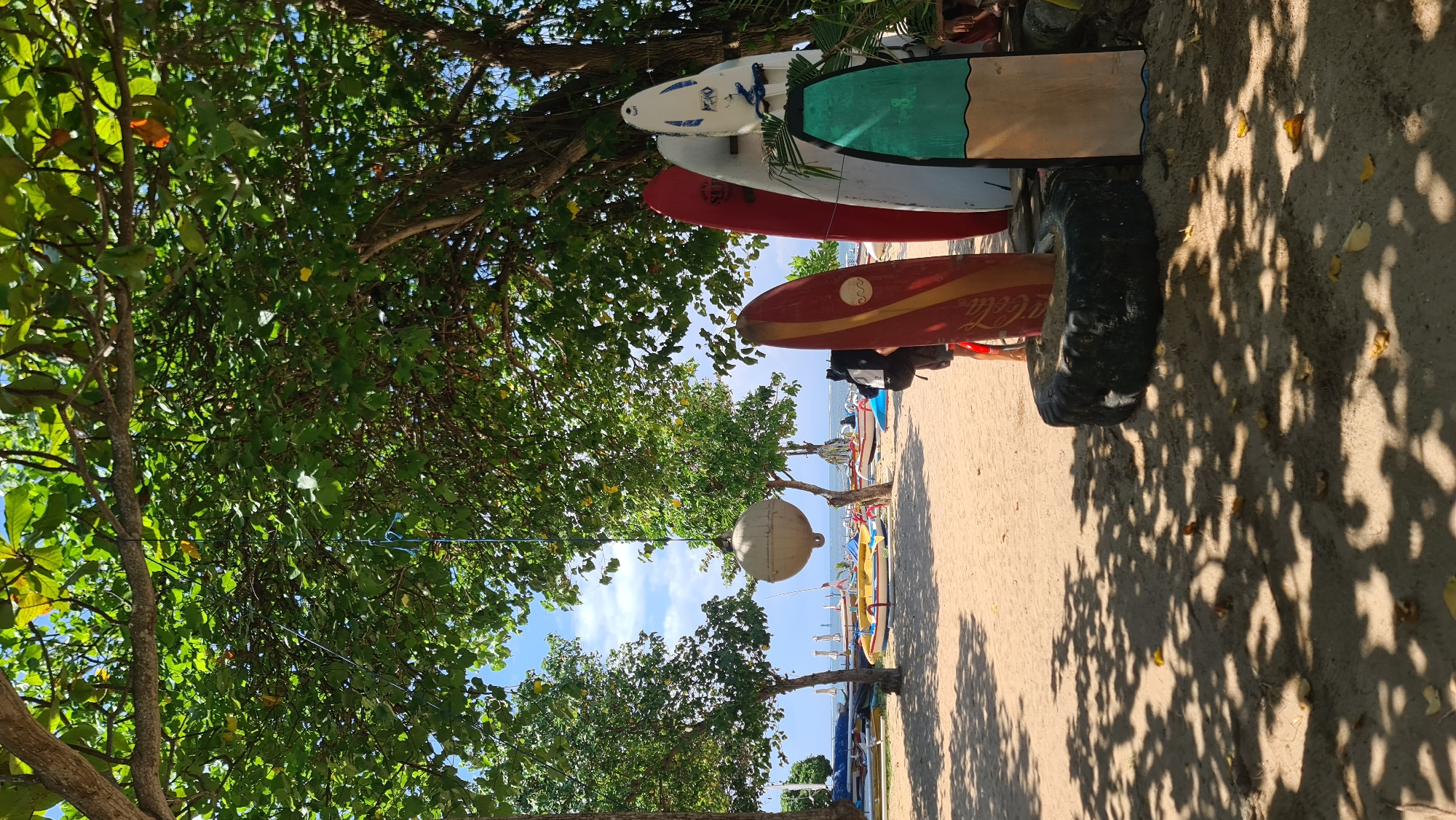 This image depicts a beach scene on a sunny day, likely in Denpasar, Indonesia. In the foreground, several surfboards are propped up against a large tire. The surfboards vary in color and design, with one featuring a red and yellow striped pattern and another with a green and tan design. Behind the surfboards, the sandy beach stretches towards the water, where several colorful boats are lined up. The boats are of a traditional Indonesian style, with outriggers. Trees with lush green foliage provide shade over parts of the beach, casting dappled shadows on the sand. In the distance, the ocean meets a bright blue sky with scattered white clouds. The overall atmosphere is one of a relaxed tropical beach destination.