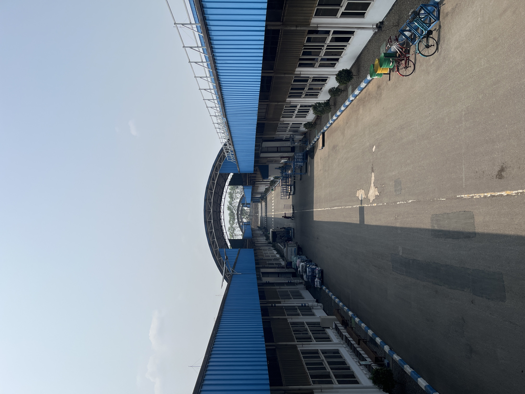 The image depicts a wide, paved road within an industrial complex, likely located in Pelang, Indonesia. On either side of the road are long, one-story buildings with blue corrugated metal roofs and white walls featuring numerous windows. These structures are connected by a series of arched, covered walkways that span the width of the road, creating a tunnel-like effect.
Several vehicles are visible on the road, including trucks and trolleys, suggesting an active industrial environment. In the foreground, on the right side of the road, are three bicycles parked in a rack, accompanied by two green and one yellow barrel. A few small trees are planted along the perimeter of the buildings.
The sky is clear and pale blue, with a few wispy clouds. The overall impression is one of an organized and functional industrial facility under a bright, sunny sky.
