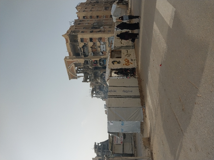 The image, taken in Khan Yunis, Palestinian Territory, depicts a street running through an area heavily affected by conflict or disaster. On the left, a row of severely damaged multi-story buildings stands, their facades torn away, exposing interiors, and structural elements crumbling. One building features a distinctive, partially ruined, arched architectural detail on its upper levels.
A series of light-colored, temporary protective barriers, possibly HESCO bastions, lines the left side of the asphalt street, separating it from the damaged structures. Several individuals are visible in the scene. Three figures, likely women dressed in black traditional clothing (abayas), are walking or standing near these barriers. Another individual in dark attire is partially obscured behind the barriers, appearing to observe the street.
The sky is clear and bright, indicating it is daytime, with shadows cast across the street suggesting the sun is relatively high. Visible text on some of the barriers includes a blue and white UNICEF logo, indicating a humanitarian presence. Other barriers display Arabic script and a stylized black and white portrait, though the text is partially obscured, it appears to contain words like "حرية" (freedom) and other messages. The scene conveys a strong sense of destruction and a challenging urban environment.