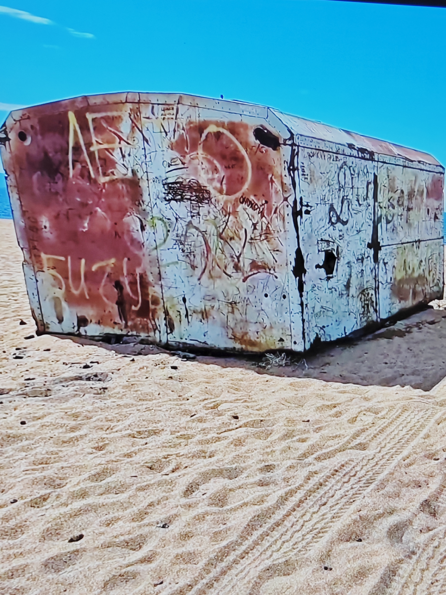 An abandoned, graffiti-covered metal structure rests on a sandy beach under a clear blue sky. The structure, possibly a shipping container or part of a larger vessel, is rectangular with a sloping top. Its surface is a chaotic canvas of spray-painted tags and markings in various colors, including red, white, yellow, and black. The paint is peeling and weathered, suggesting exposure to the elements. In the foreground, the sand is imprinted with tire tracks, indicating recent vehicle activity. The ocean is visible in the background, appearing as a thin blue line. The overall scene conveys a sense of desolation and decay.