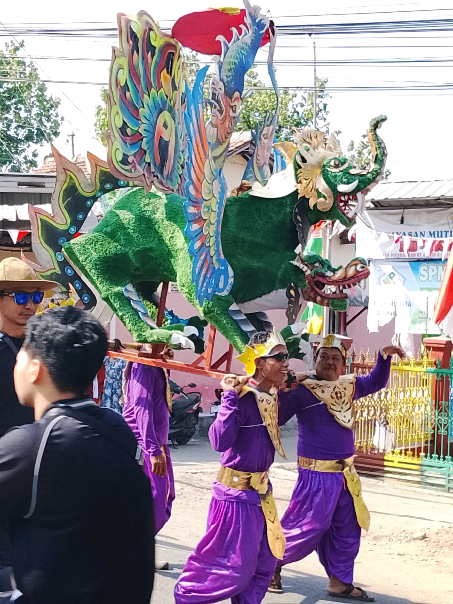 In Ujungrusi, Indonesia, an outdoor street scene captures a parade or procession featuring a large, colorful dragon effigy. The effigy, predominantly green with vibrant blue, red, yellow, and black accents, is carried on a wooden structure by several individuals. Two men in matching bright purple outfits with gold trim and headwear are prominent, walking and holding poles that support the effigy. Another person in a purple outfit is visible further back. In the foreground, a person wearing a black shirt and a straw hat with sunglasses is looking towards the effigy. The background shows buildings with red roofs, trees, and a brightly colored fence. Electrical wires are visible overhead, and a motorcycle is parked on the side of the road. The overall atmosphere suggests a festive cultural event.