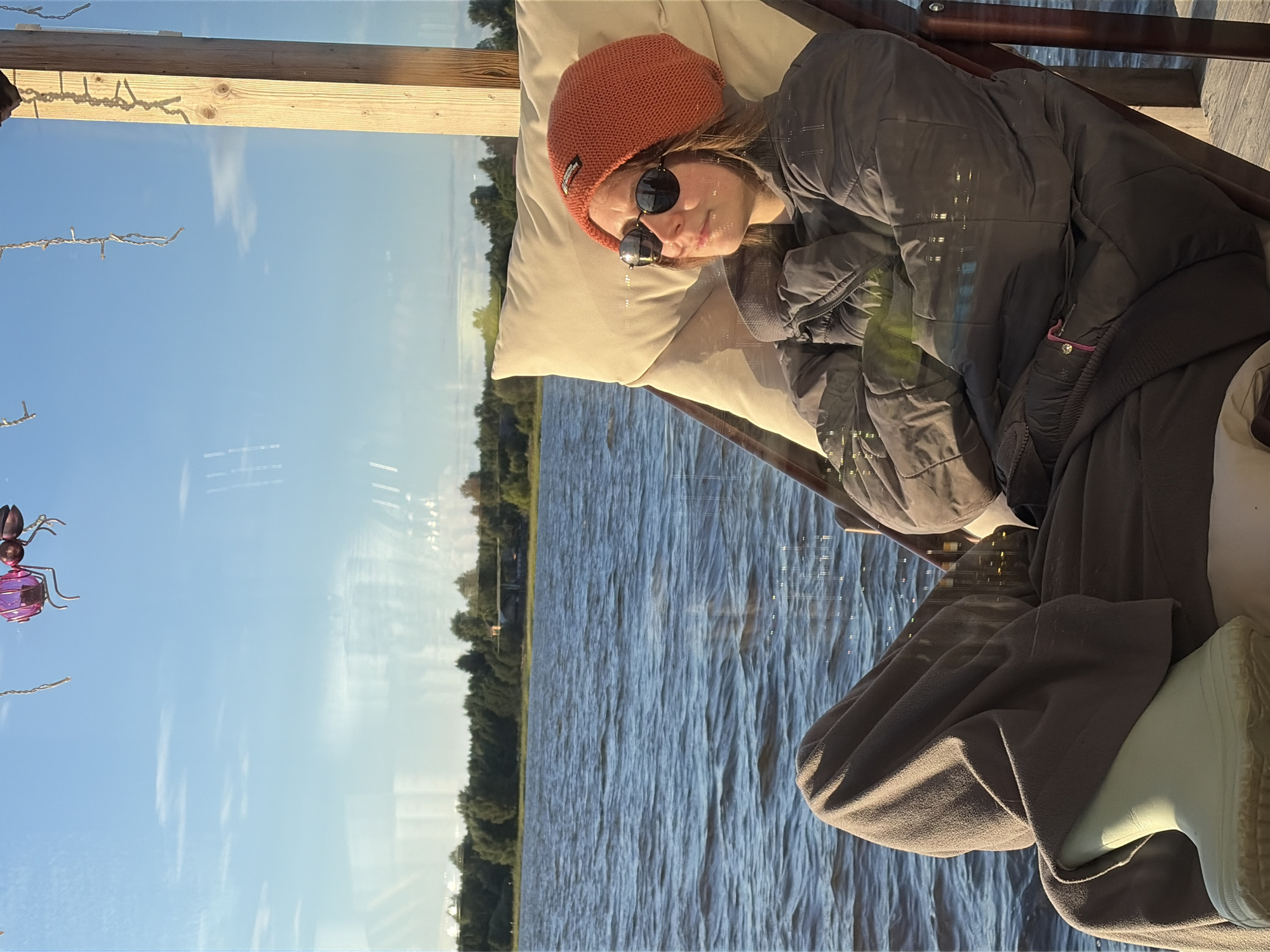 A young woman sits in a reclined chair on a wooden dock, looking out at a body of water. She is wearing a grey puffer jacket, brown pants, a bright orange beanie, and sunglasses. Her legs are covered by a grey blanket, and she is wearing light green rain boots. The water is calm with small ripples, and a tree line with houses and buildings is visible in the distance. The sky is blue with scattered white clouds. A wooden post is visible to the right of the woman, and decorative string lights and a pink insect-shaped ornament hang from above, suggesting a whimsical outdoor setting, potentially a recreational area or private residence in the city of Vidogoshchi, Russia, near the Volkhov River. The lighting indicates it is late afternoon or early evening.