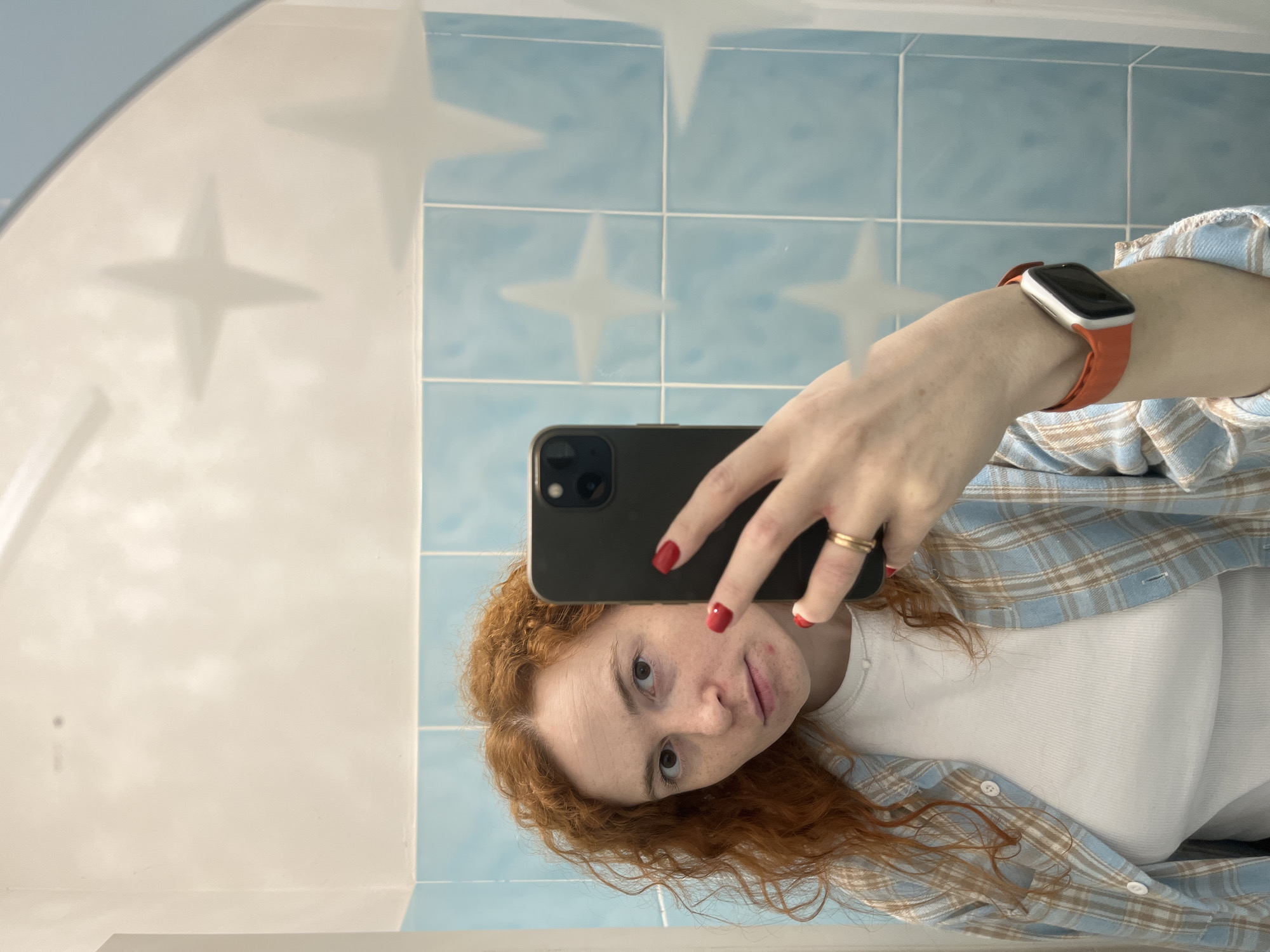 A person with red curly hair is taking a selfie in a bathroom. The person is wearing a white t-shirt and a light blue plaid shirt. They are holding a black smartphone in their right hand. The person is also wearing an orange smartwatch on their left wrist and a gold ring on their left ring finger. The bathroom has light blue tiles with white stars. The wall is white. The location is Chaikovsky, Russia.