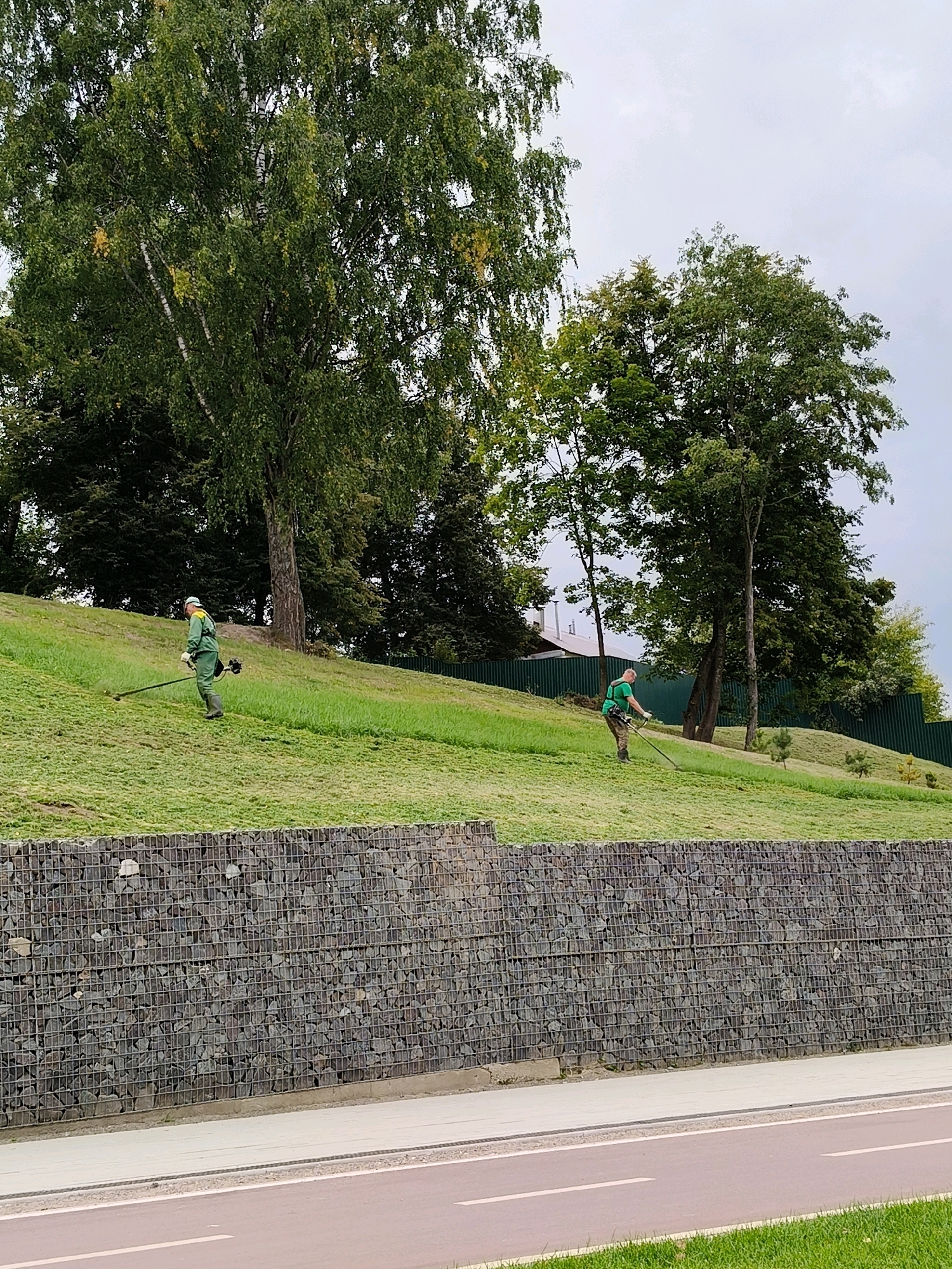 Two individuals, dressed in work attire, are engaged in mowing a grassy slope. The person on the left is wearing a green jumpsuit and boots, operating a string trimmer. The person on the right is wearing a green shirt and pants, also with a string trimmer. The slope is steep and covered in green grass, with several trees visible in the background. A gabion wall, constructed with wire cages filled with stones, forms a retaining wall at the base of the slope. In the foreground, a paved pathway or road is visible with painted lines. The sky above is overcast. The scene appears to be an outdoor public space, possibly a park or roadside in Naro-Fominsk, Russia.