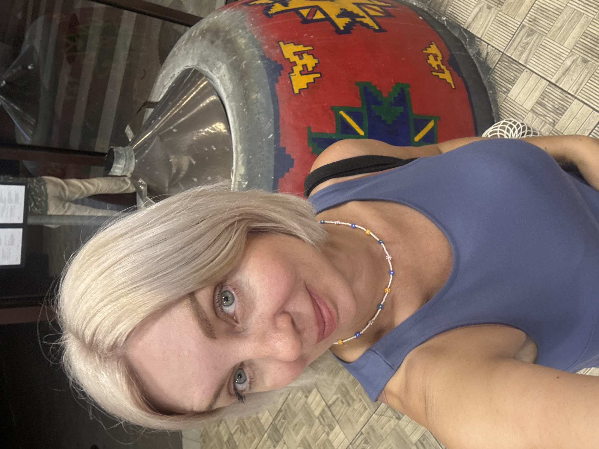 A woman with short, blonde hair and blue eyes is taking a selfie in front of a large, round, red object decorated with colorful geometric patterns. She is wearing a blue tank top and a beaded necklace. The object appears to be a tandoor oven, a traditional clay oven used for cooking. In the background, a person is visible wearing light-colored pants. The floor is tiled with a pattern of grey and white rectangles. The location is identified as Sirius, Russia.