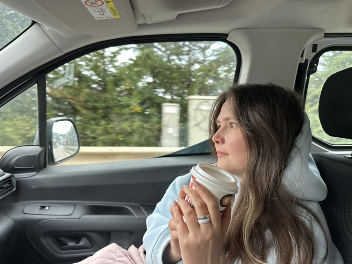 An adult female is observed in the front passenger seat of a vehicle, looking towards her right out the side window. She has long, dark brown hair and is wearing a light blue hooded sweatshirt over light pink trousers. Her left hand holds a white disposable coffee cup with red and brown logo markings, and a white band-style ring is on her left ring finger. The vehicle's interior consists of black door paneling, with a side mirror visible through the window. An "AIRBAG" warning label is affixed to the overhead visor. Outside the vehicle, green foliage, likely trees, and a beige road barrier are present. Water droplets are discernible on the exterior of the window glass. The scene is situated in Bartula, Montenegro.