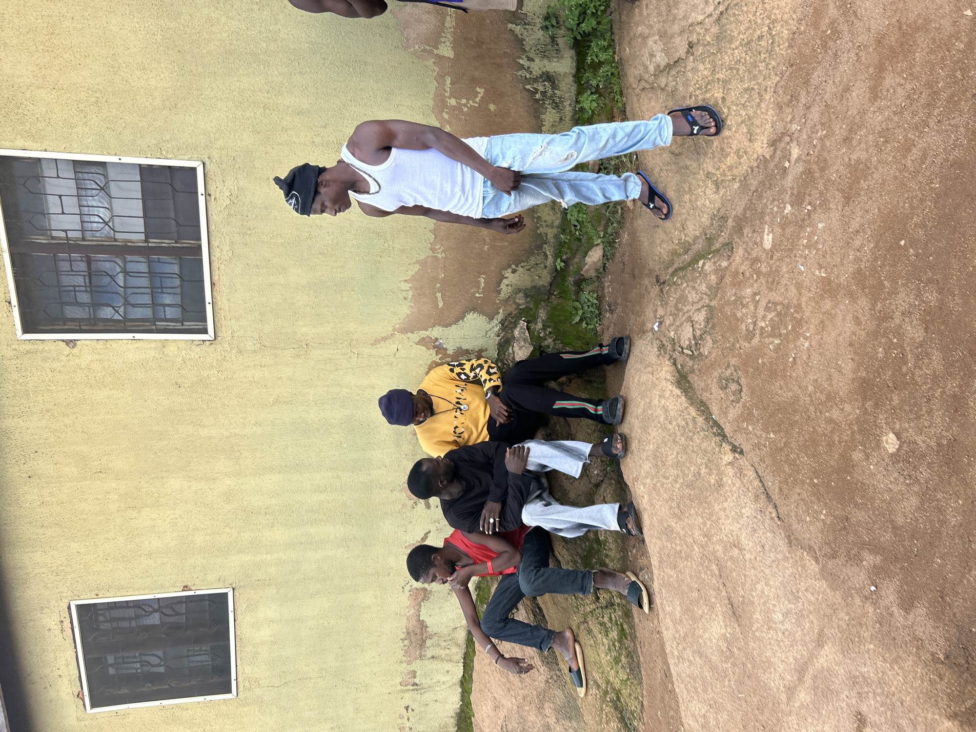 Four young men are gathered outside a weathered, pale yellow building. The building has two windows with protective bars and a textured facade showing signs of age and water damage. The ground in front of the building is a mix of rough, reddish-brown earth and large, flat rocks.
On the rocks, three men are seated. The man on the far left wears a red t-shirt and dark jeans, with his arm resting on his knee and his chin propped on his hand. He has on dark sandals. Next to him, a man in a black long-sleeved shirt and light gray pants looks towards the right. He is wearing dark sandals and has a ring on his finger. The third seated man, in the middle, wears a yellow long-sleeved shirt with a black and yellow patterned design on the sleeves and a dark beanie. He appears to be looking down and interacting with the man next to him.
Standing to the right, a man in a white tank top and ripped light blue jeans is looking to his left. He is wearing black flip-flops and has a silver chain around his neck. A portion of a fifth person, wearing a dark shirt, is visible on the far right edge of the frame.
The scene is consistent with an outdoor environment in the city of Jos, Nigeria, characterized by the building's architecture, the ground material, and the casual attire of the individuals.