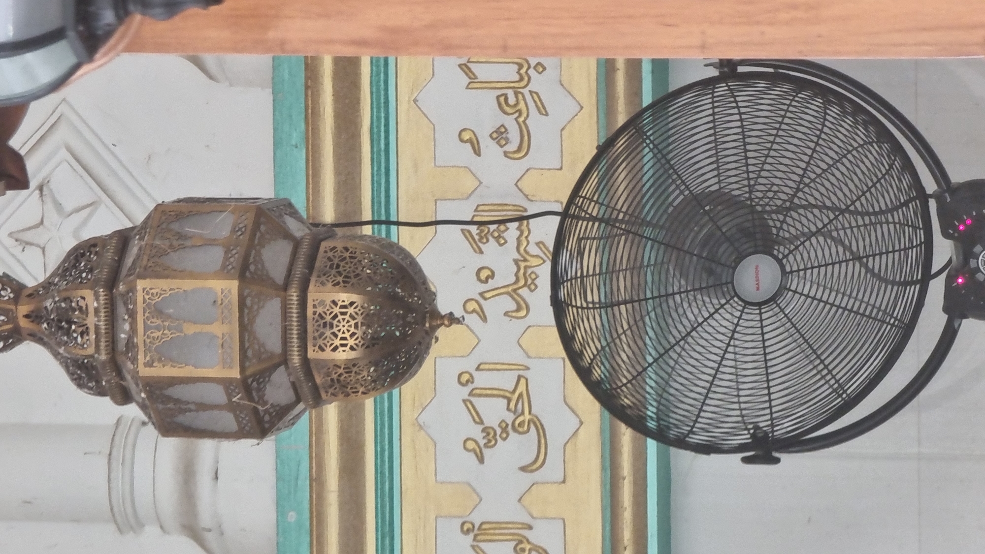 The image shows an ornate, brass-colored lantern hanging above a black oscillating fan in what appears to be an indoor setting. The lantern has intricate geometric patterns and is illuminated by light filtering through its panels. Below the lantern, a wall is adorned with golden Arabic calligraphy set against white star-shaped motifs. The fan, positioned in the foreground, is industrial in design with a wire grill and a central logo that reads "MASION". The background consists of a white wall with architectural elements. This scene is located in Lapang Timu, Indonesia, likely within a mosque or a similarly decorated public building given the calligraphy and architectural style.