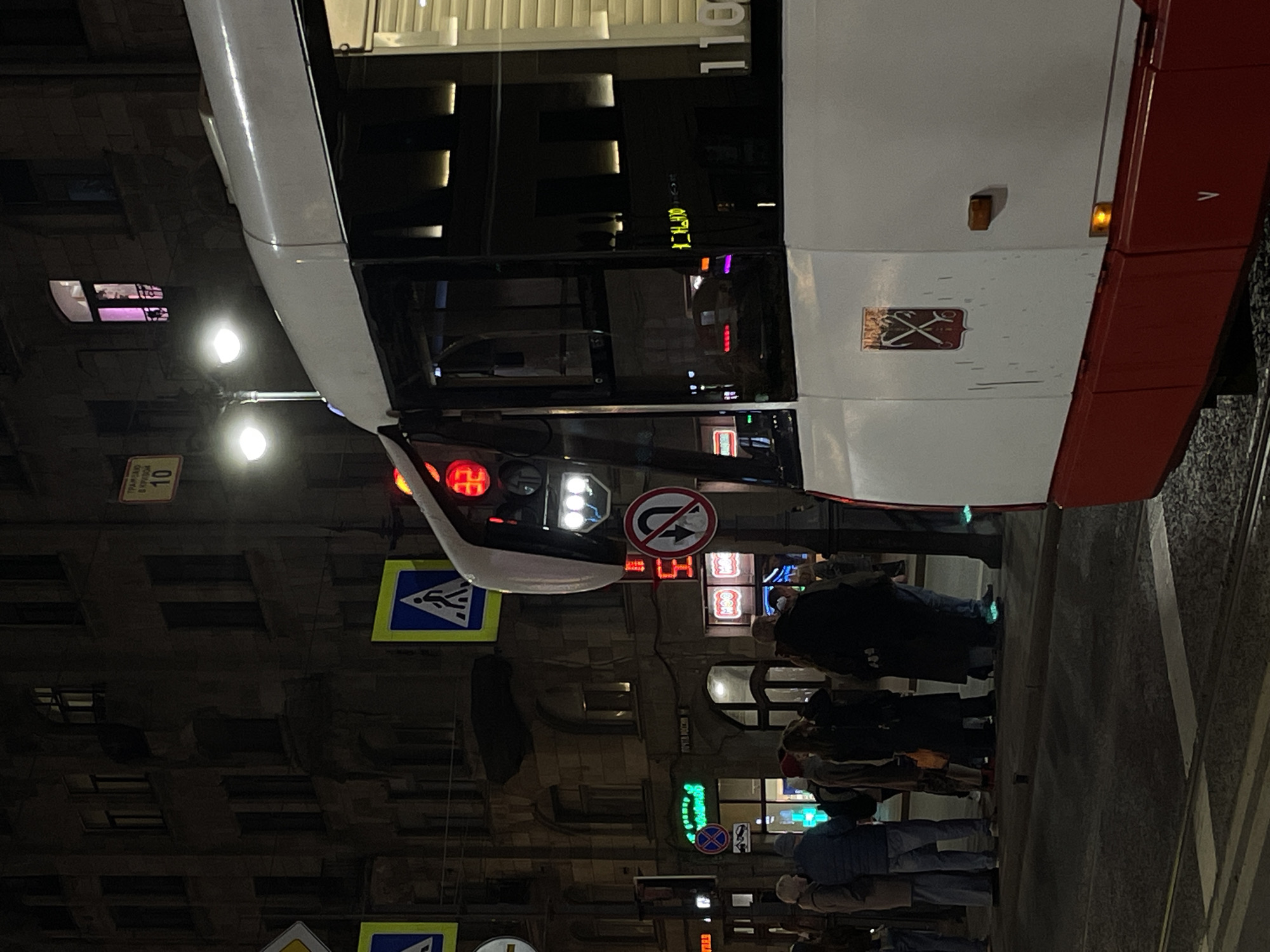 The image captures an urban scene in Saint Petersburg, Russia, at night. A tram, predominantly white with a red stripe on the right side, is in the foreground, obscuring a partial view of a building. The building across the street has windows illuminated with a soft, warm light. Pedestrians are walking on the sidewalk. Several street signs are visible, including pedestrian crossing signs and a "no U-turn" sign. Neon signs and streetlights contribute to the nighttime illumination. The overall scene conveys a bustling urban atmosphere.