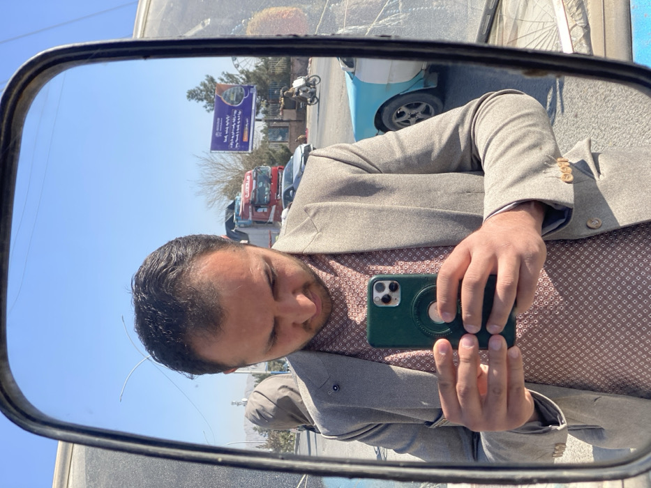 A man in a gray suit and patterned shirt takes a selfie in a side-view mirror. His phone has a green case. The mirror reflects a sunny street scene with trucks, a blue car, and a billboard in the background. The angle suggests he is in a vehicle.